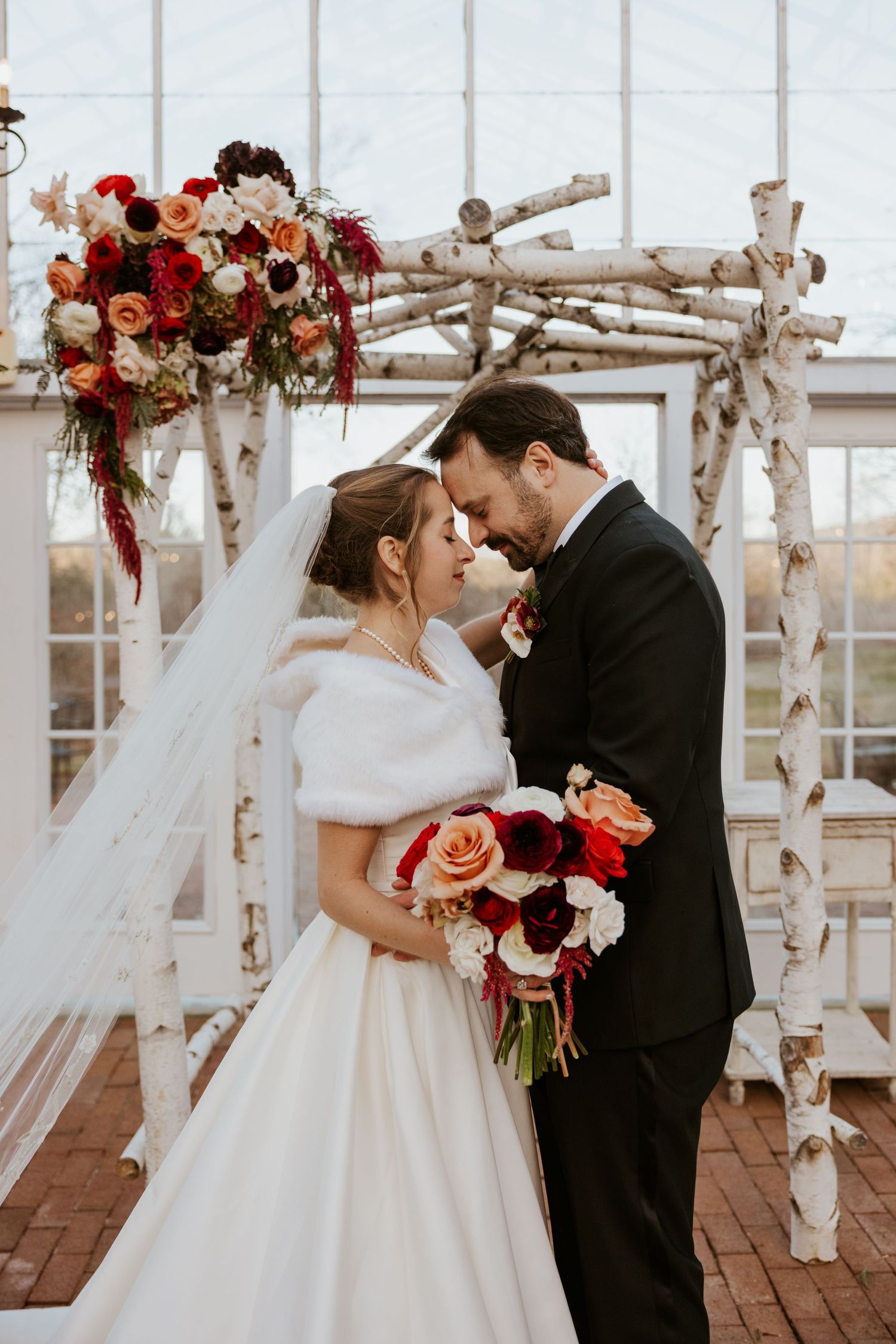 Bride and groom share intimate moment at birch wood altar with red and white floral arrangements.