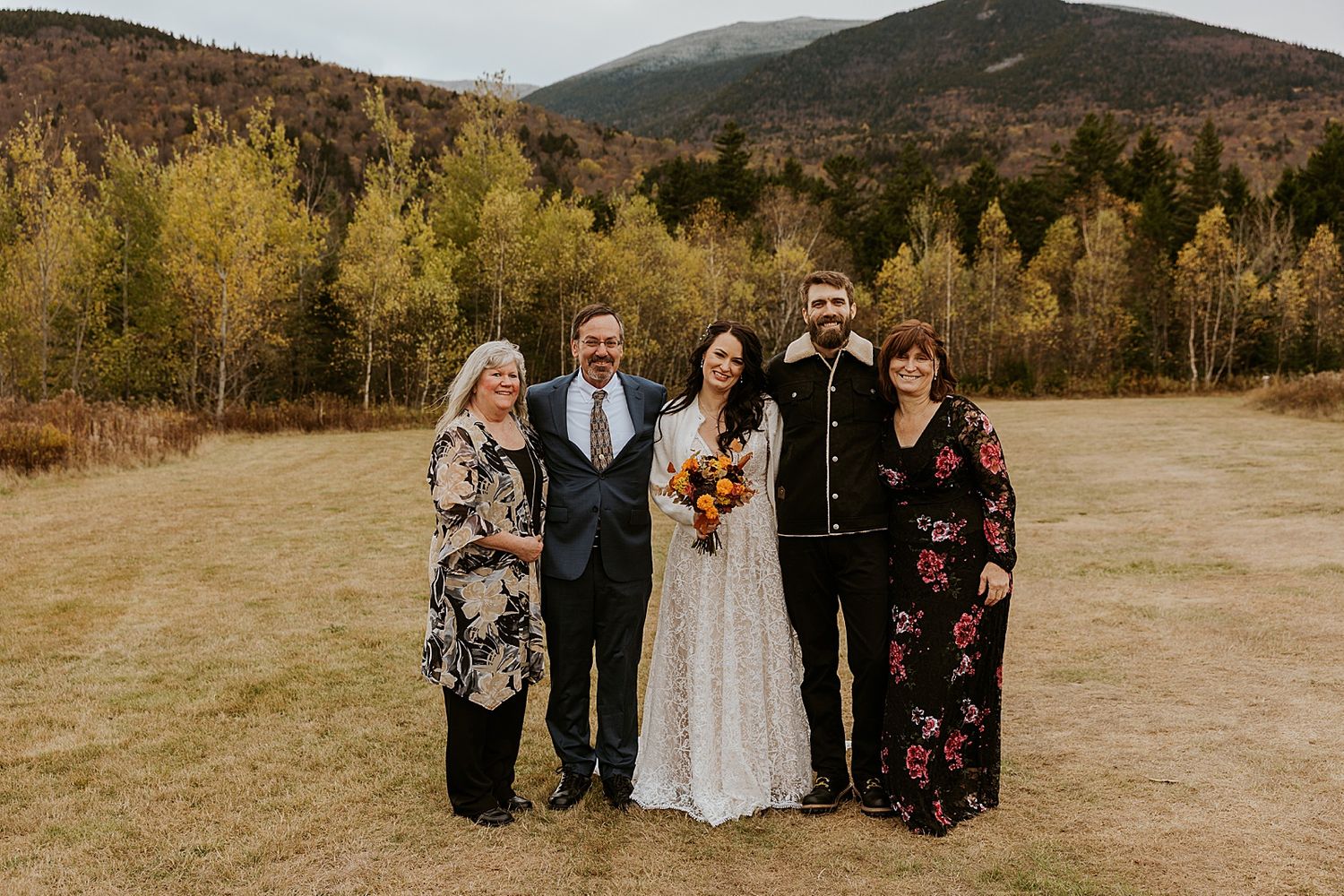Wedding party poses outdoors in autumn mountain meadow, bride in lace gown with bouquet flanked by family.