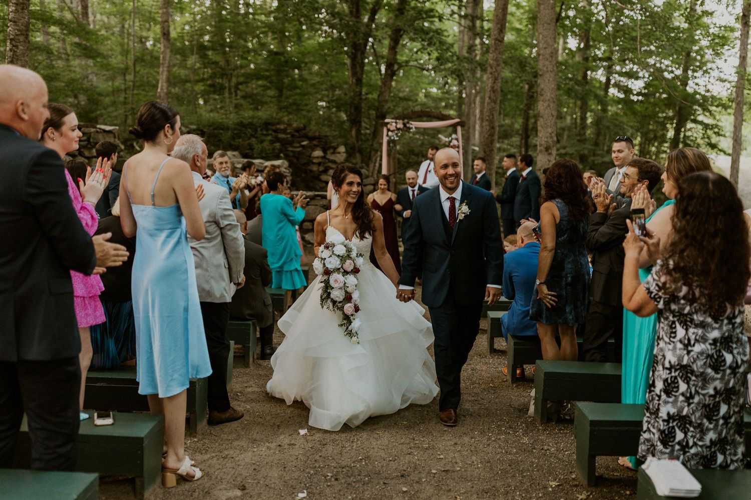 Wedding celebration in a rustic outdoor forest venue with guests gathered.