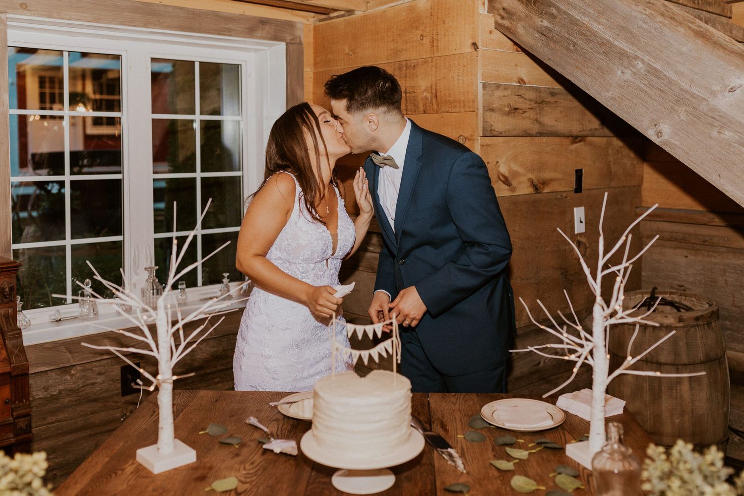 Couple sharing a sweet moment while cutting their white tiered wedding cake at a rustic venue.