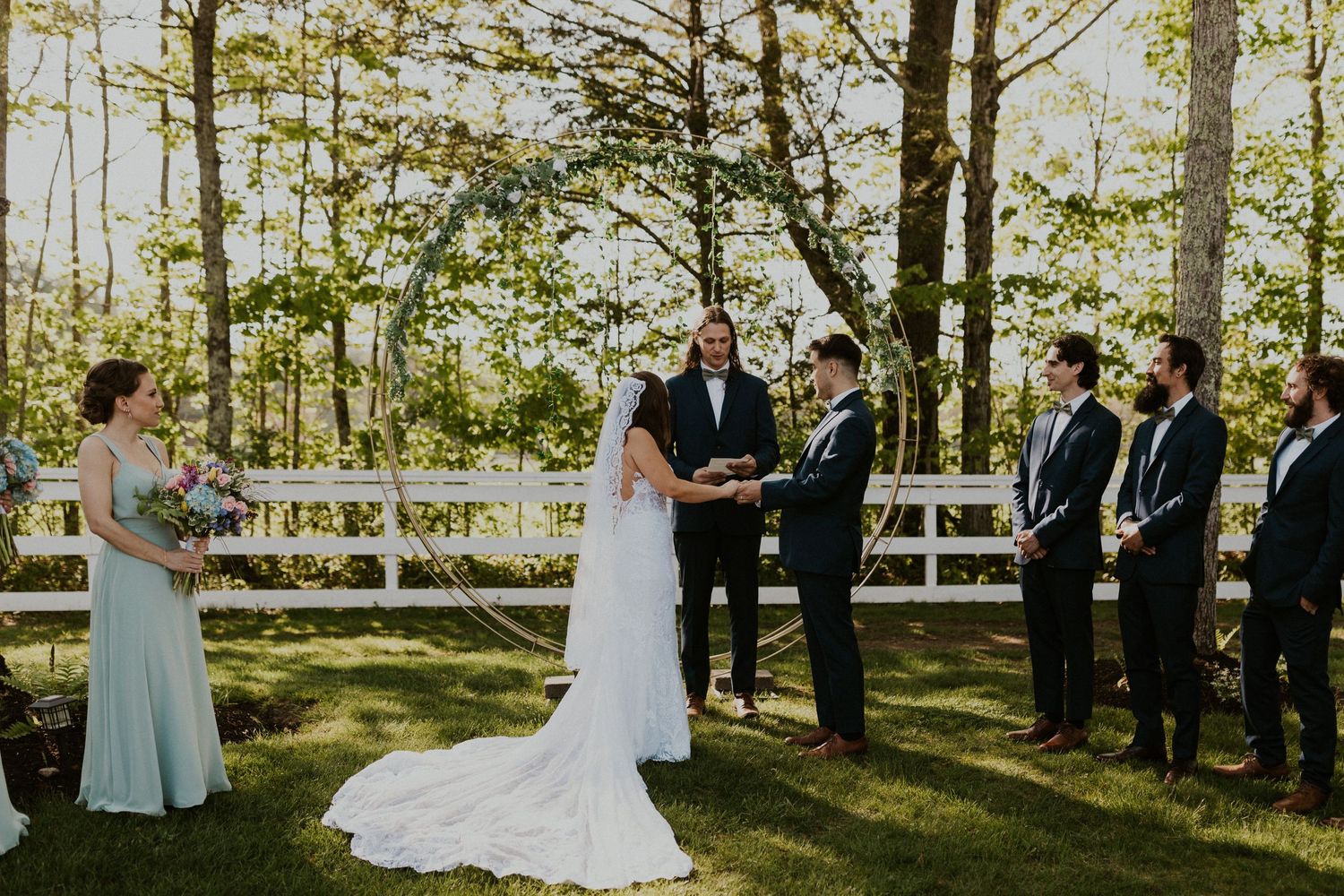 Chrissy and Mike holding hands as their wedding ceremony begins. It is a lovely spring day with the sun shining.