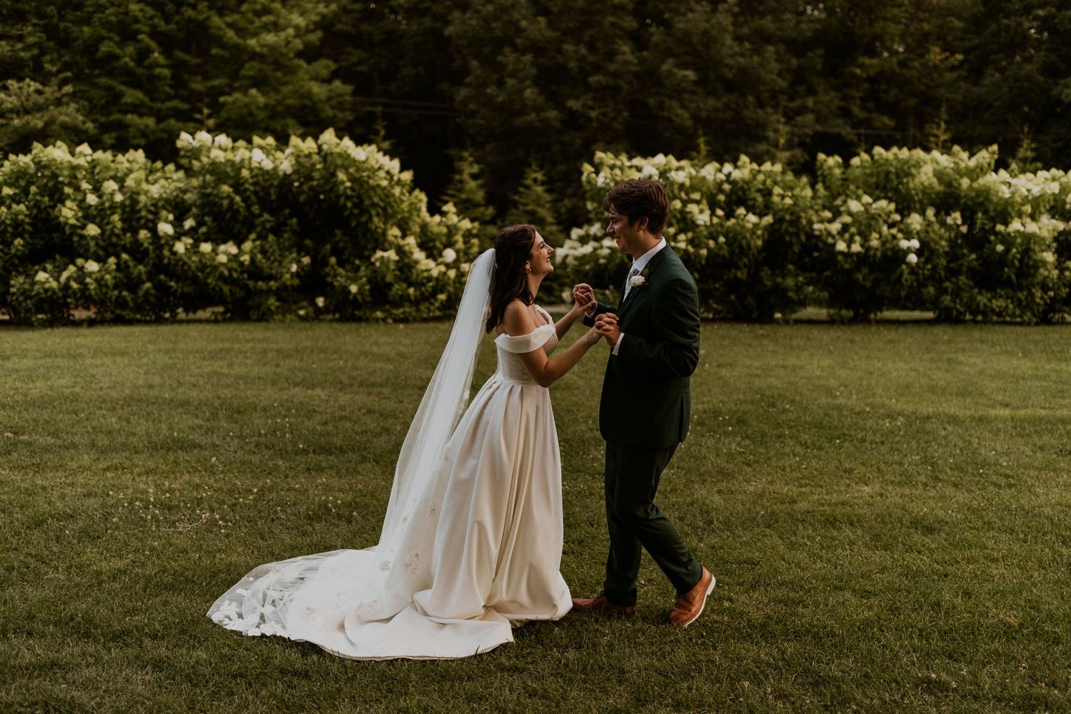 A romantic wedding couple dancing together on a lush lawn with blooming white hydrangea bushes in the background.