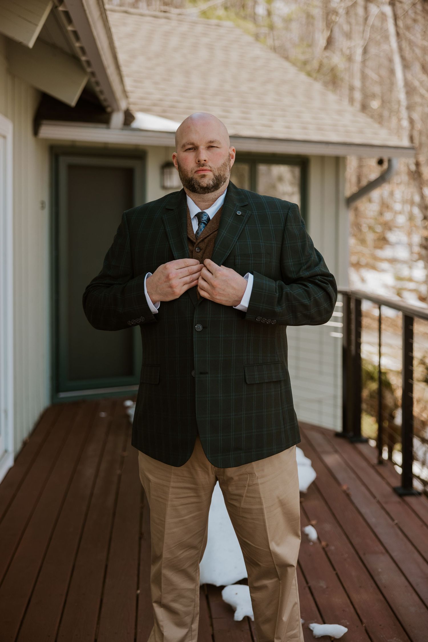Groom adjusts his tie while standing on a wooden deck porch wearing a dark suit jacket and khaki pants.