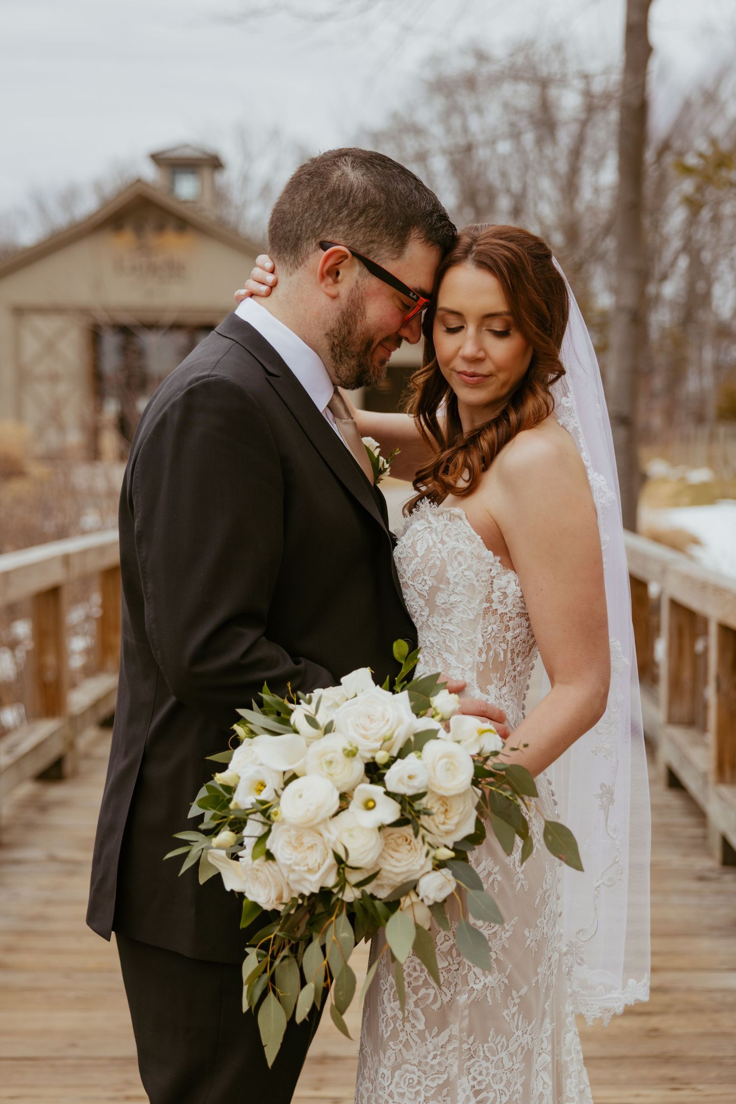 A romantic wedding couple embraces on a wooden bridge while holding an elegant white floral bouquet.