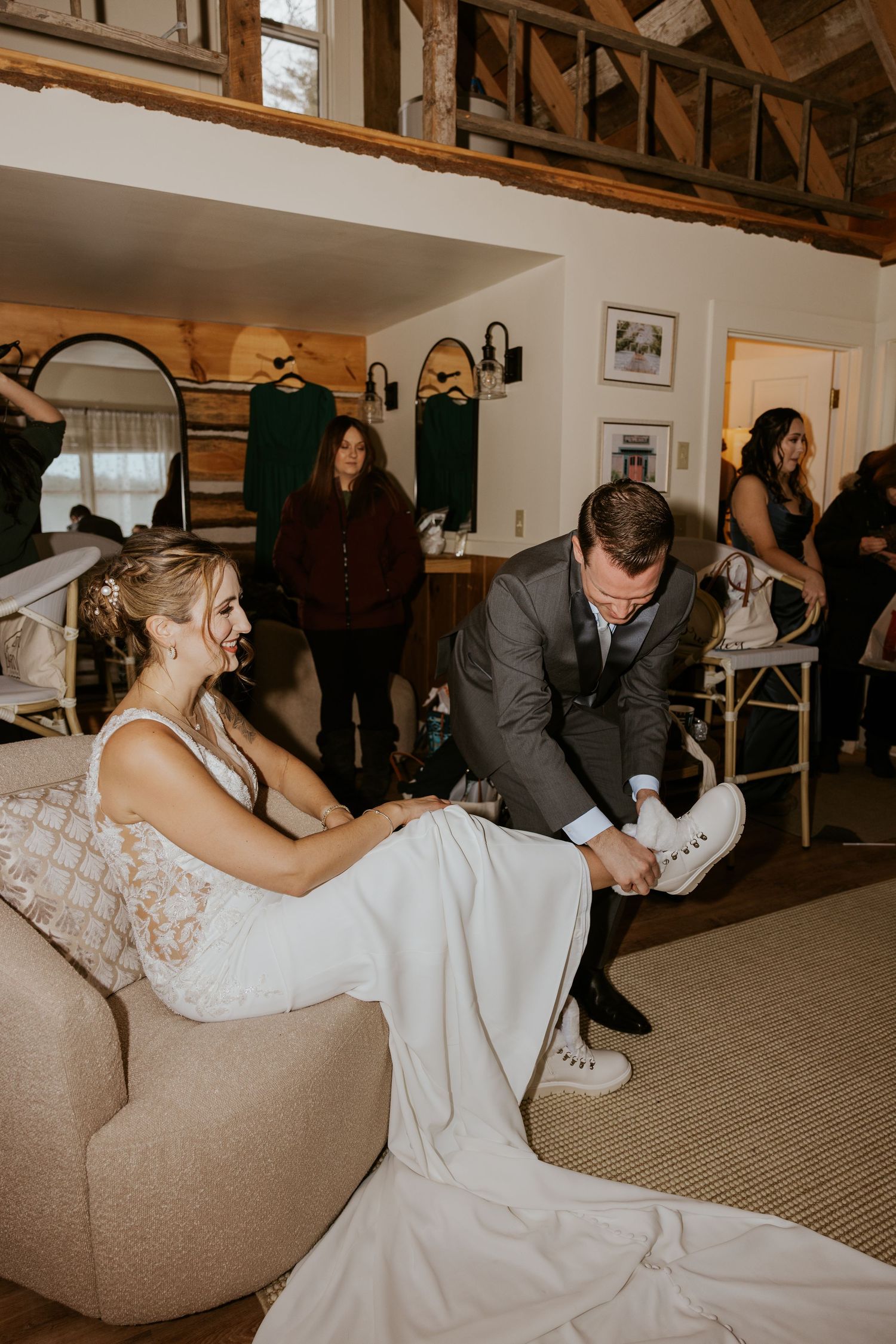 Groom helping bride put on white sneakers while she smiles in wedding dress on a couch at rustic barn venue.