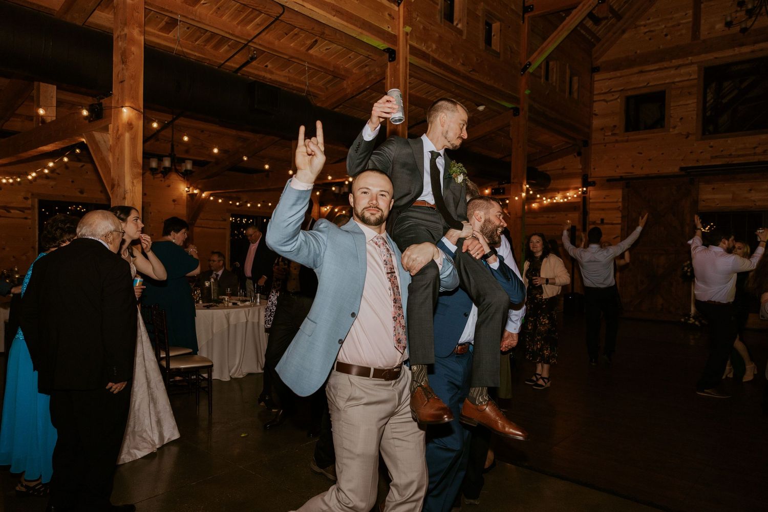 Wedding reception guests celebrate and dance joyfully inside a rustic barn venue decorated with string lights.
