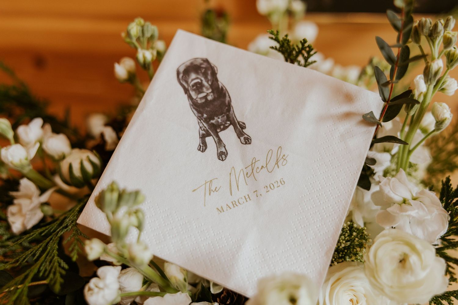A watercolor illustration of a dog decorates a wedding invitation card among white floral arrangements.