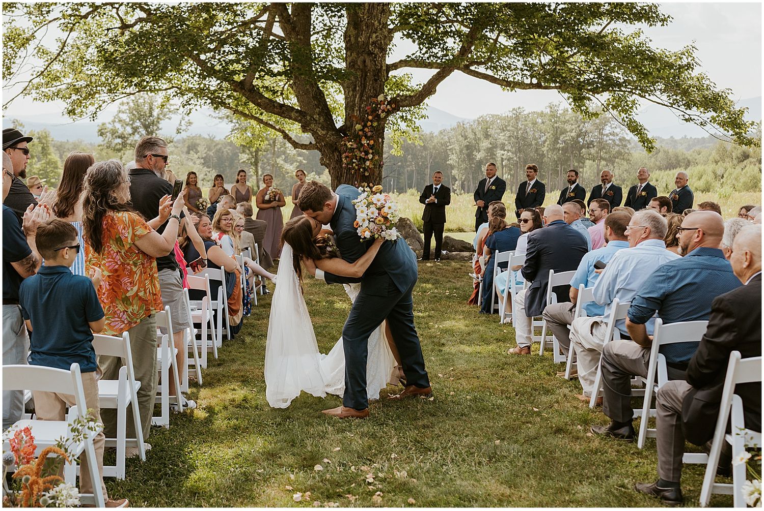 Outdoor wedding ceremony setup with white chairs arranged under sprawling tree.