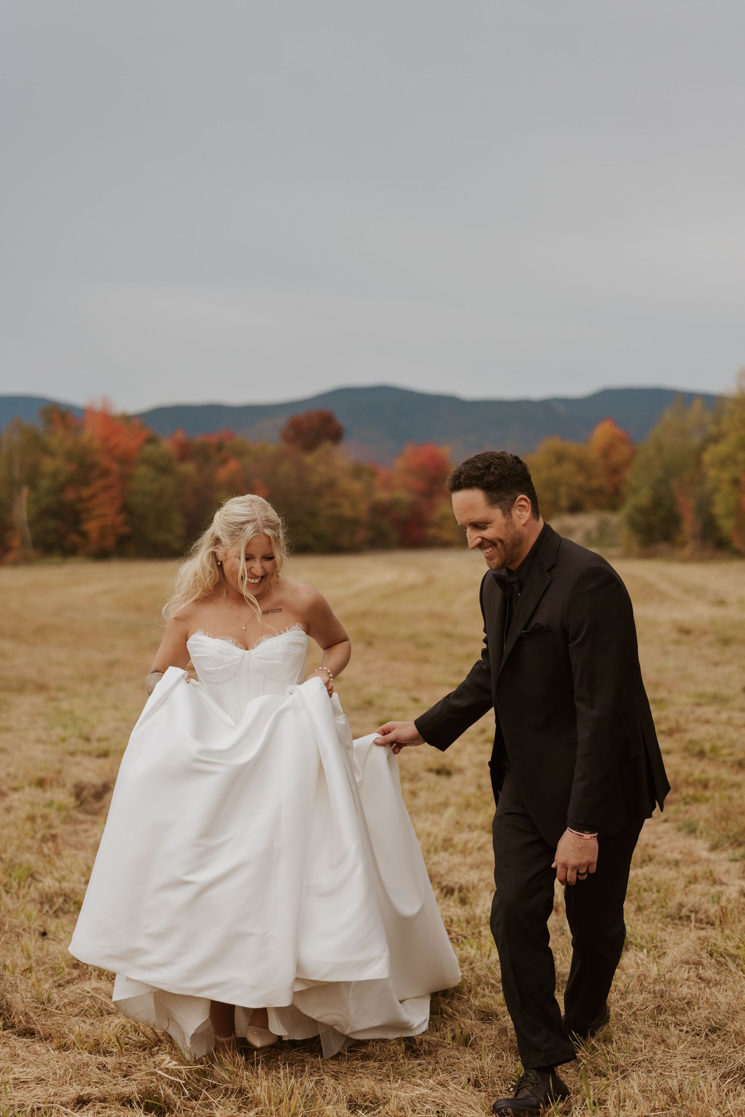 Bride and groom walk together through golden field with vibrant fall foliage and mountains in background.