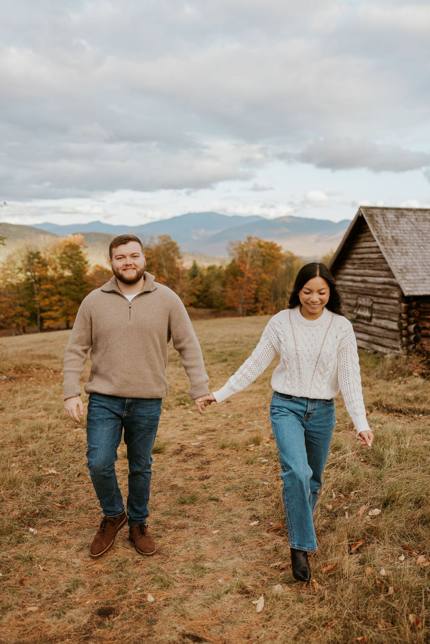Smiling couple holding hands walking in an open field with mountain views.
