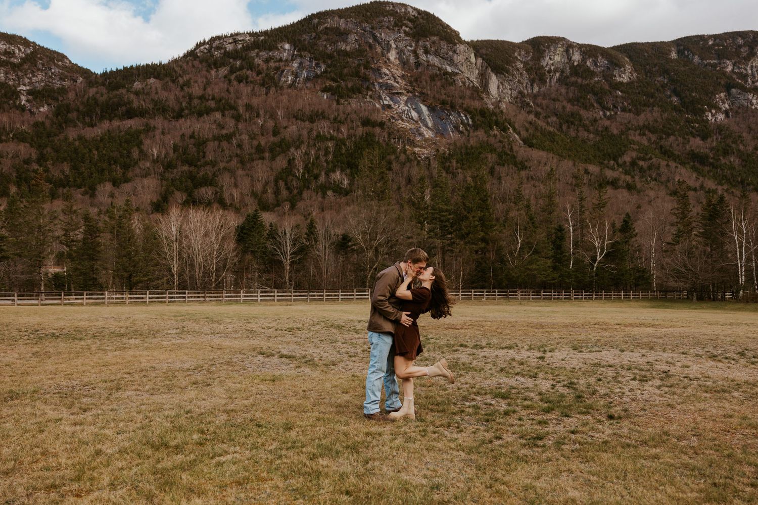 Man lifting and embracing woman in a grassy mountain field with rocky cliffs and evergreen trees in the background.