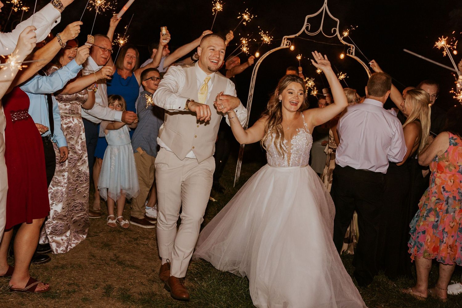Bride and groom exit through sparkler tunnel surrounded by cheering wedding guests at night.