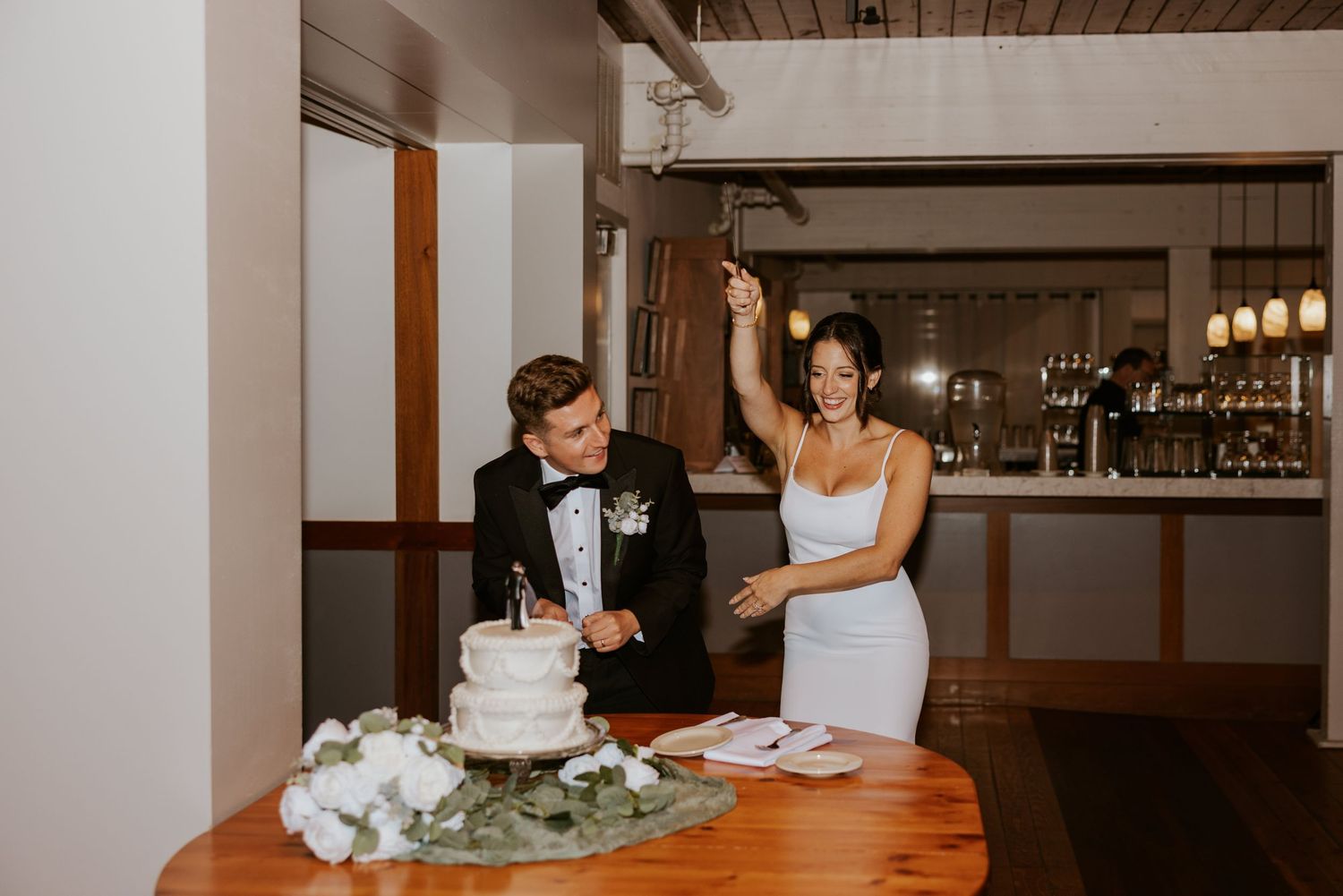 Bride celebrating while groom cuts tiered white wedding cake at reception.