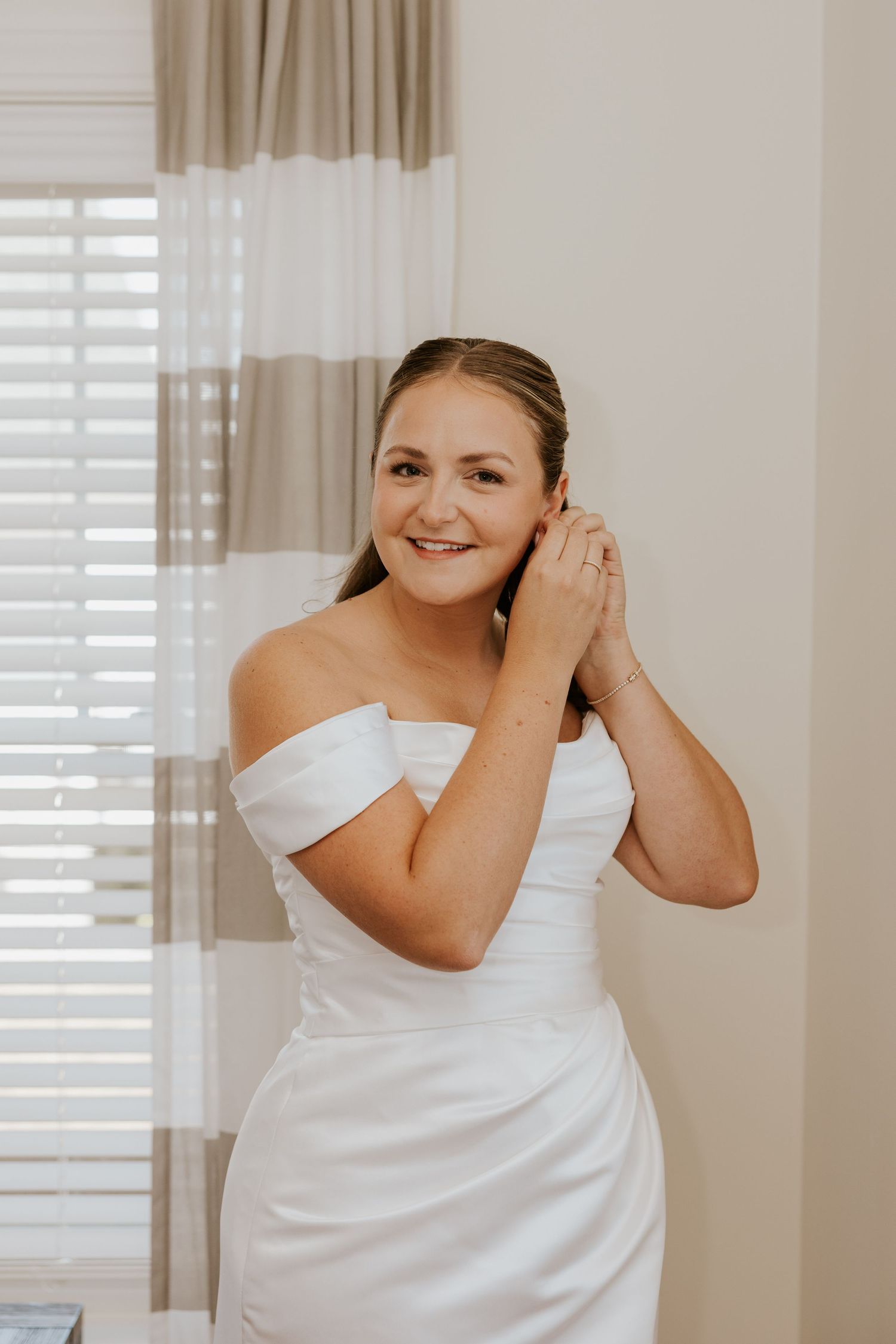 Smiling bride in white off-shoulder wedding gown putting on earrings while standing near a bright window.