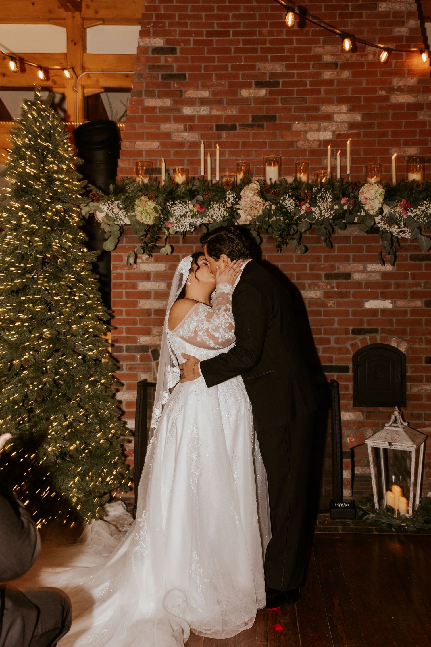 Bride and groom sharing first kiss beside a decorated Christmas tree at a winter wedding ceremony.