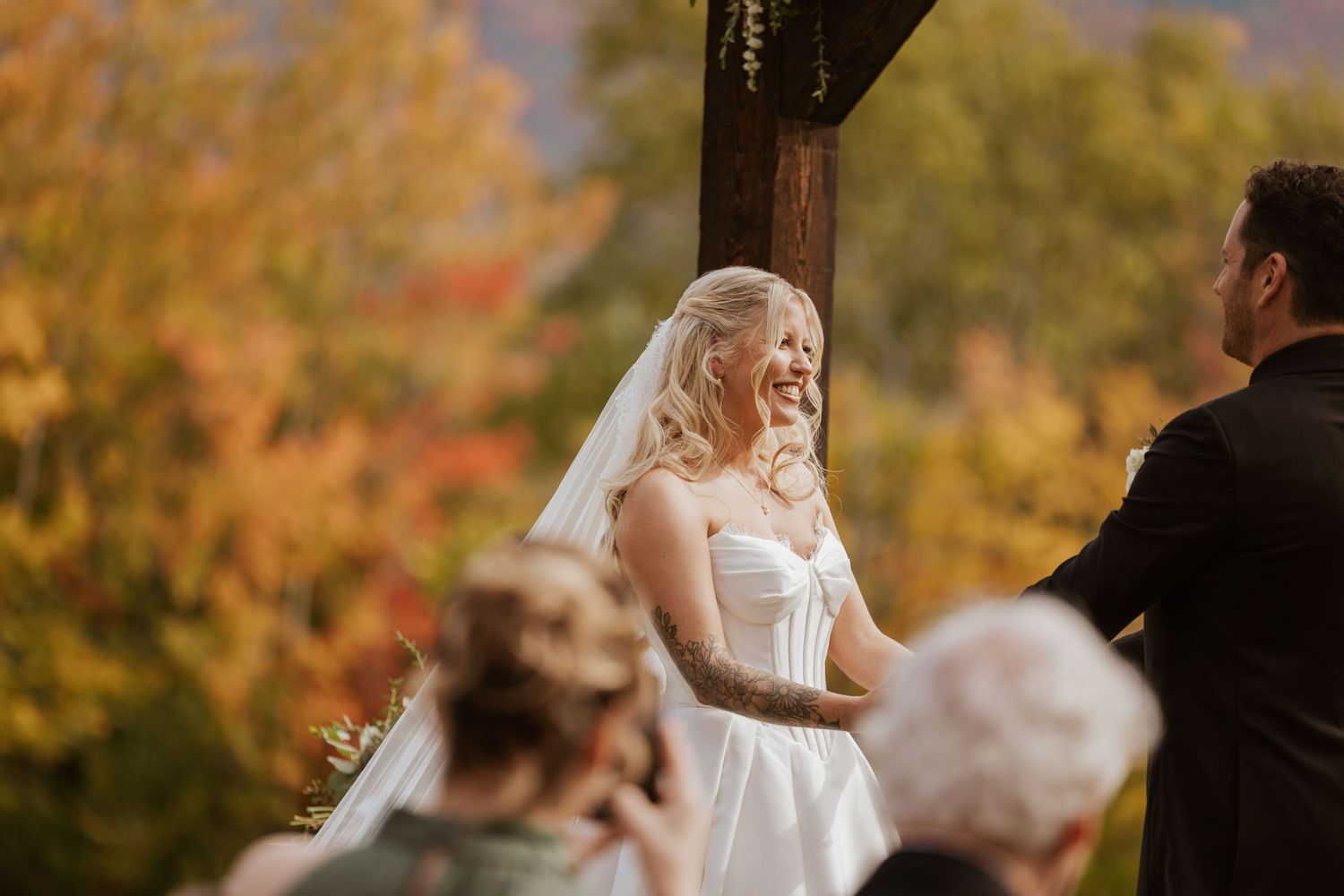 Smiling bride in white strapless gown and veil at outdoor fall wedding ceremony with colorful autumn foliage backdrop.