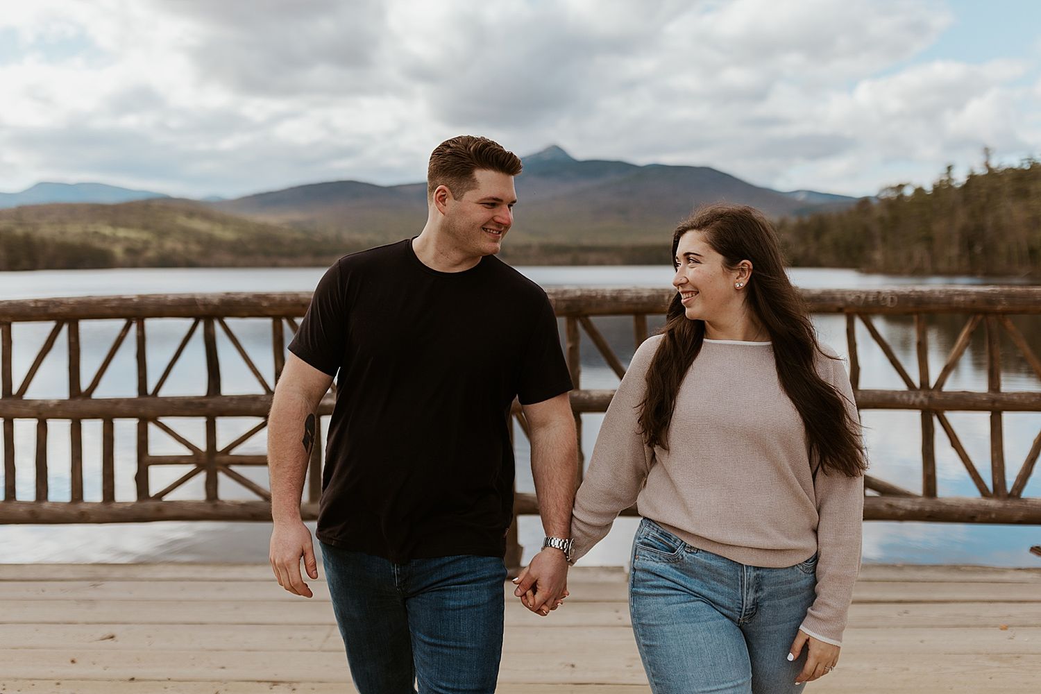 Happy couple holding hands on wooden bridge with mountain lake backdrop on cloudy day.