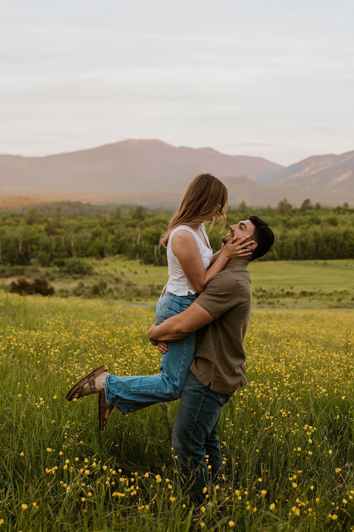 Man lifting woman in a flower field with mountains in the background during a romantic outdoor couples photo session.