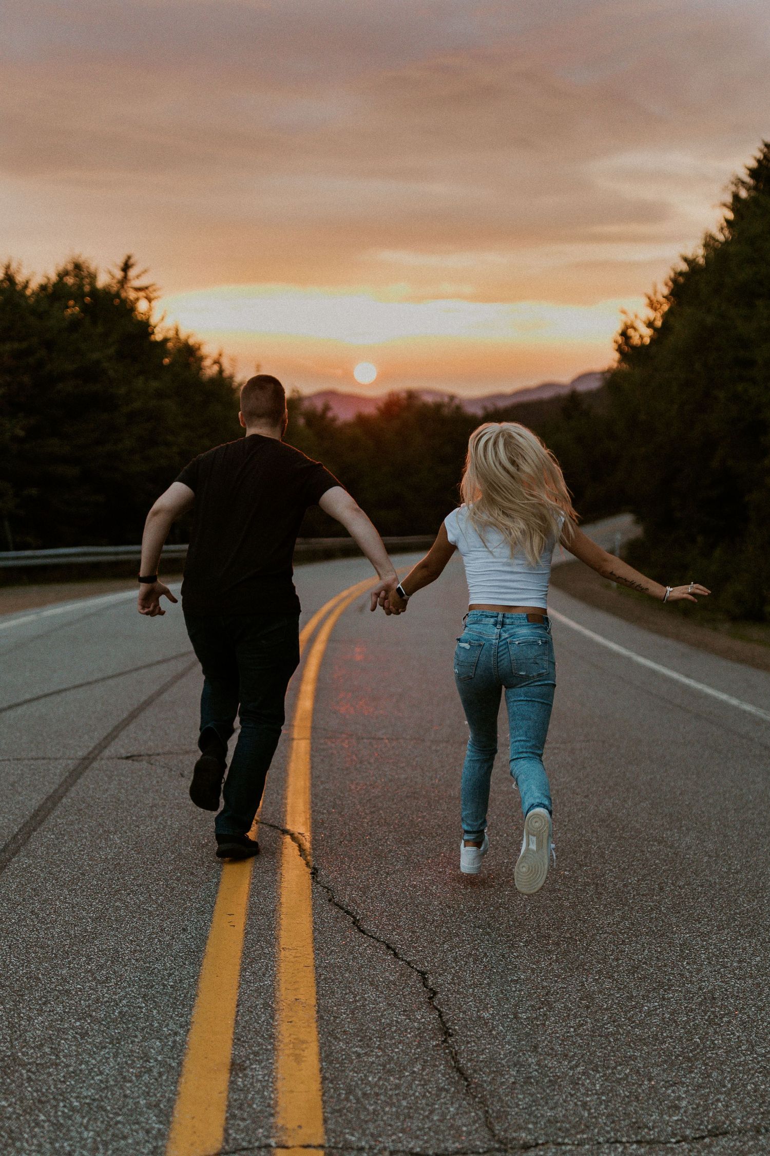 Paige and Justin holding hands and running down the Kancamagus Highway in Lincoln NH playfully together at sunset.