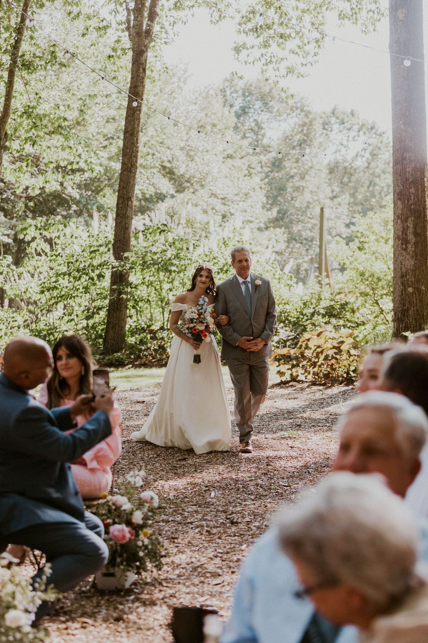 Outdoor forest wedding ceremony with guests seated on wooden benches among tall trees.