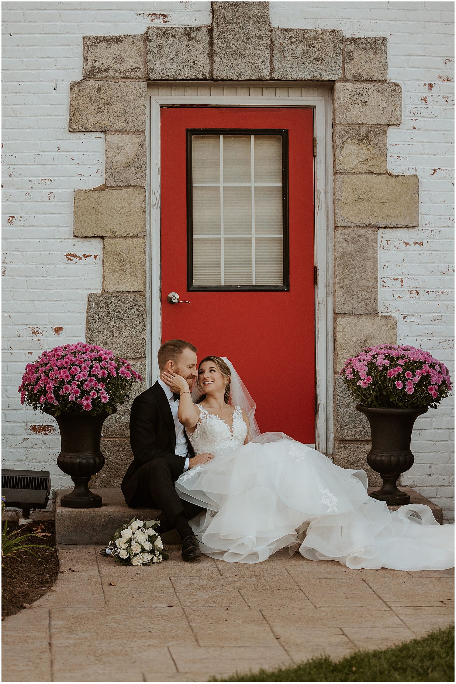 Wedding couple sitting by a red door with pink flowers in stone planters.