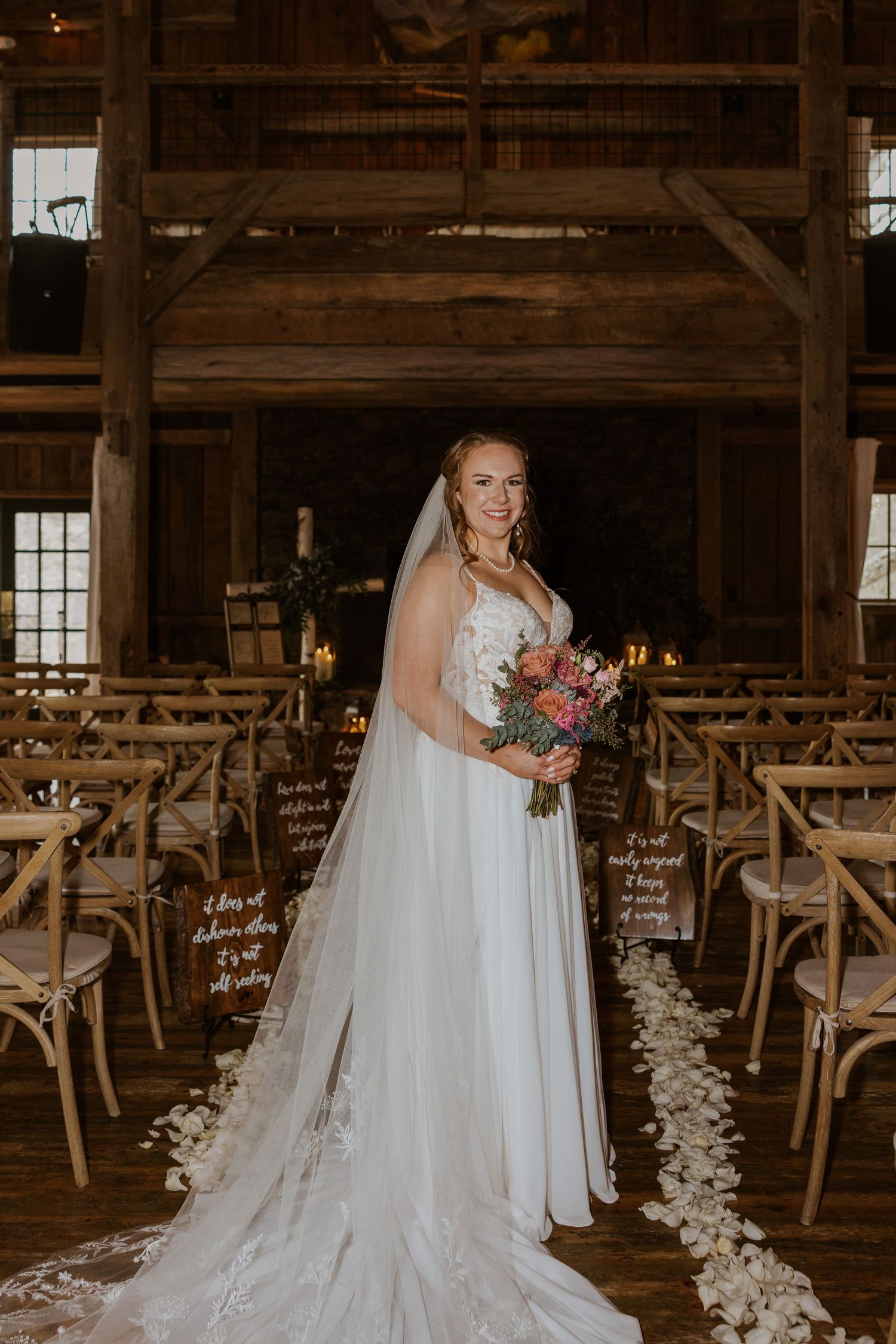 Bride in flowing white gown posing in rustic barn venue with floral bouquet.