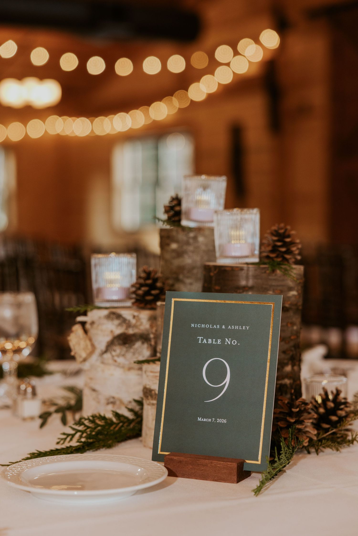 Rustic wedding table decor with pinecones, birch logs and table number 9 under warm string lights.