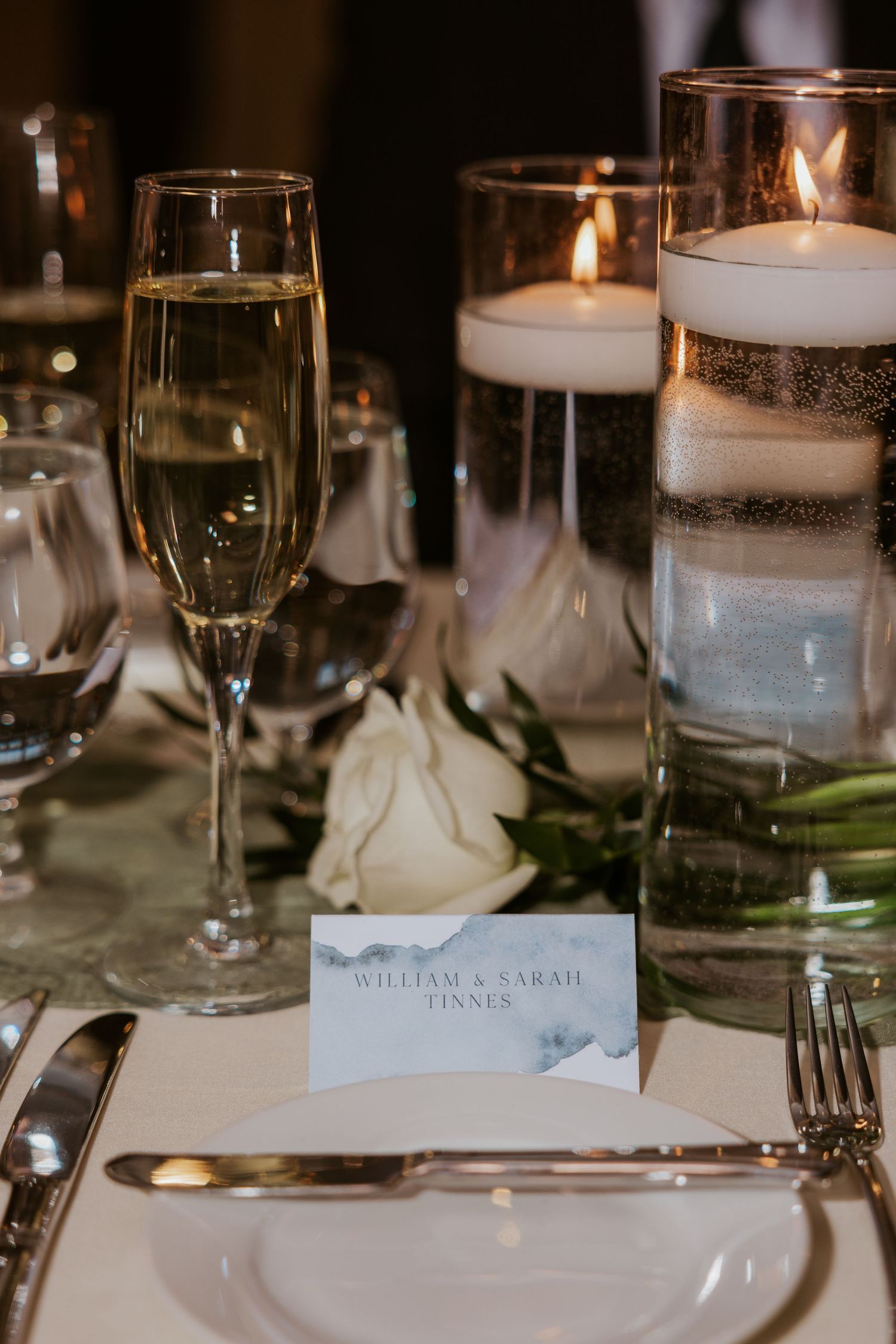 Wedding reception table setting with elegant glassware, candles and place cards in moody lighting.