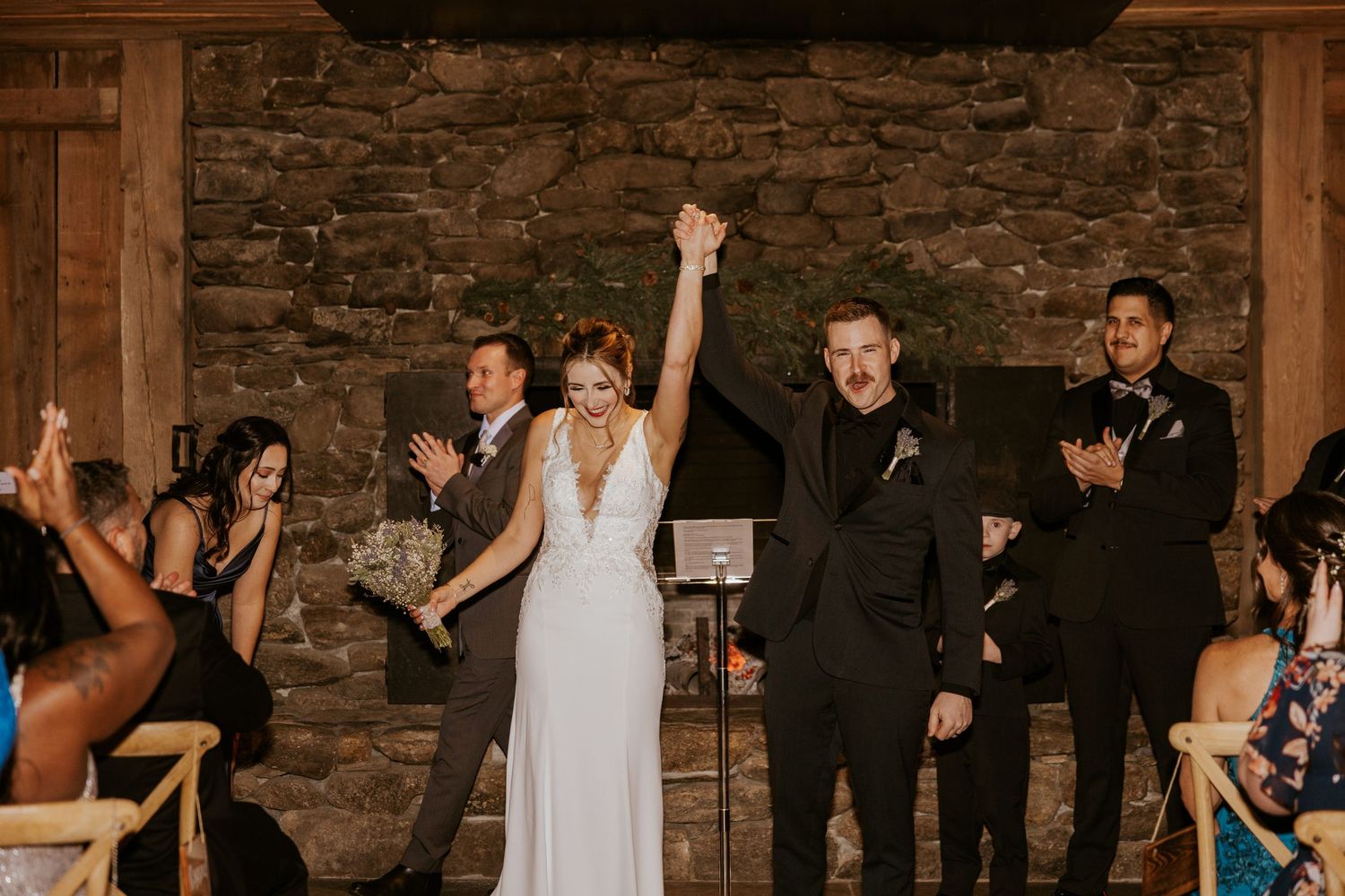 Newlyweds raising hands in celebration at rustic stone wall wedding ceremony as guests applaud.