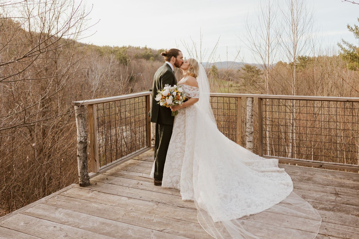 Bride and groom share romantic kiss on wooden deck overlook surrounded by autumn trees and mountains.