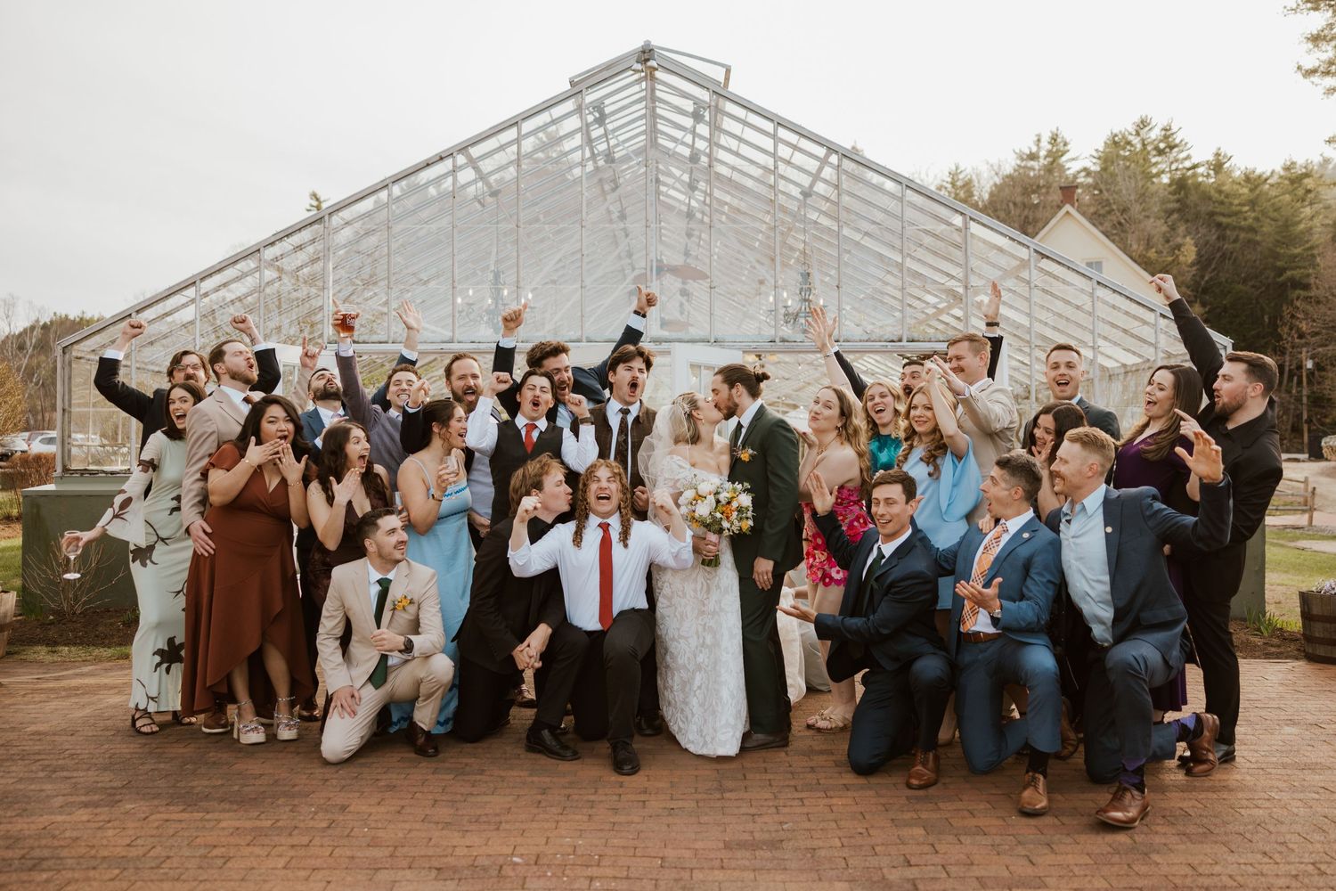 Joyful wedding party celebrating in front of a glass greenhouse, bride and groom surrounded by guests raising glasses.