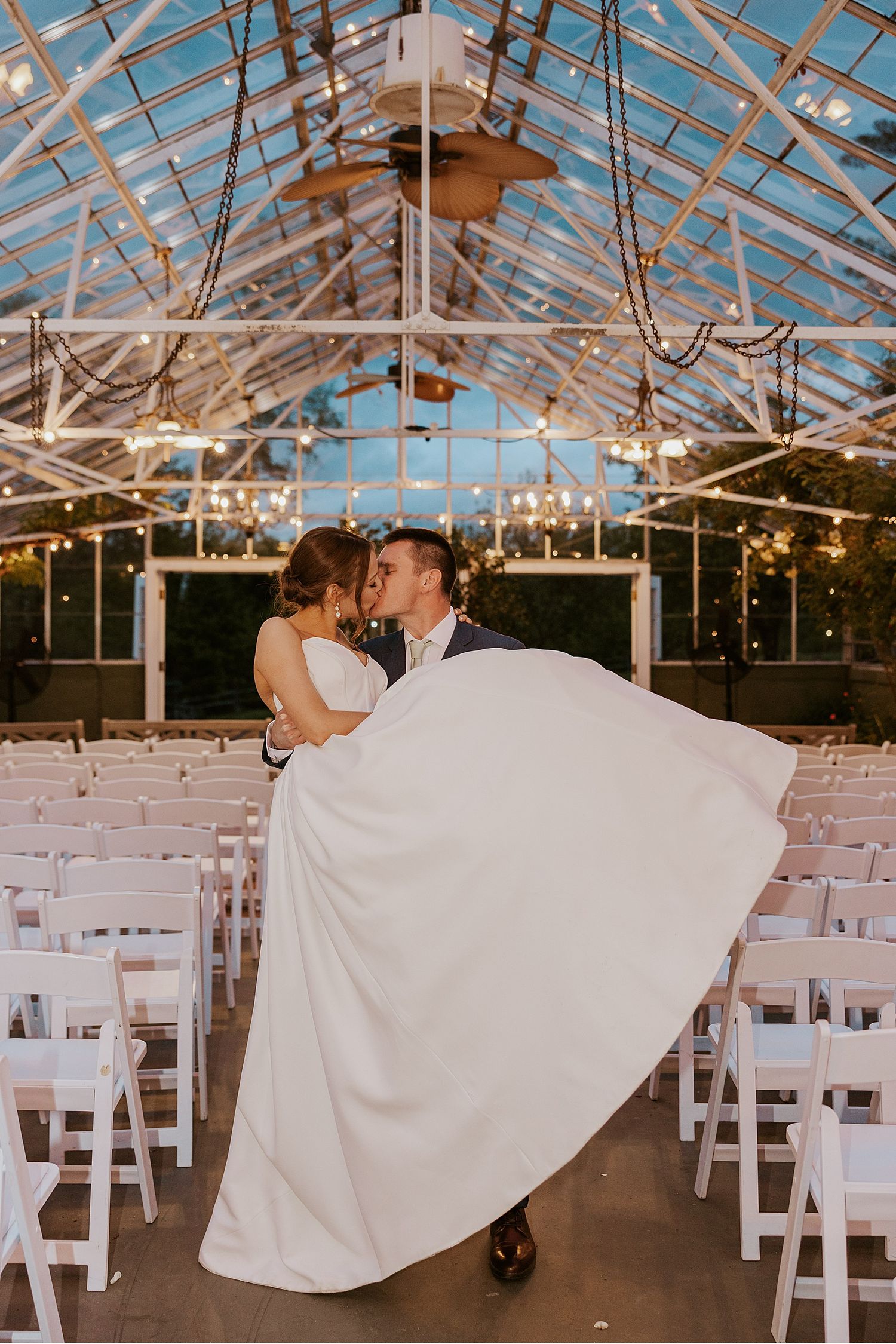 Glass greenhouse wedding ceremony setup with white chairs and string lights creating a magical atmosphere.