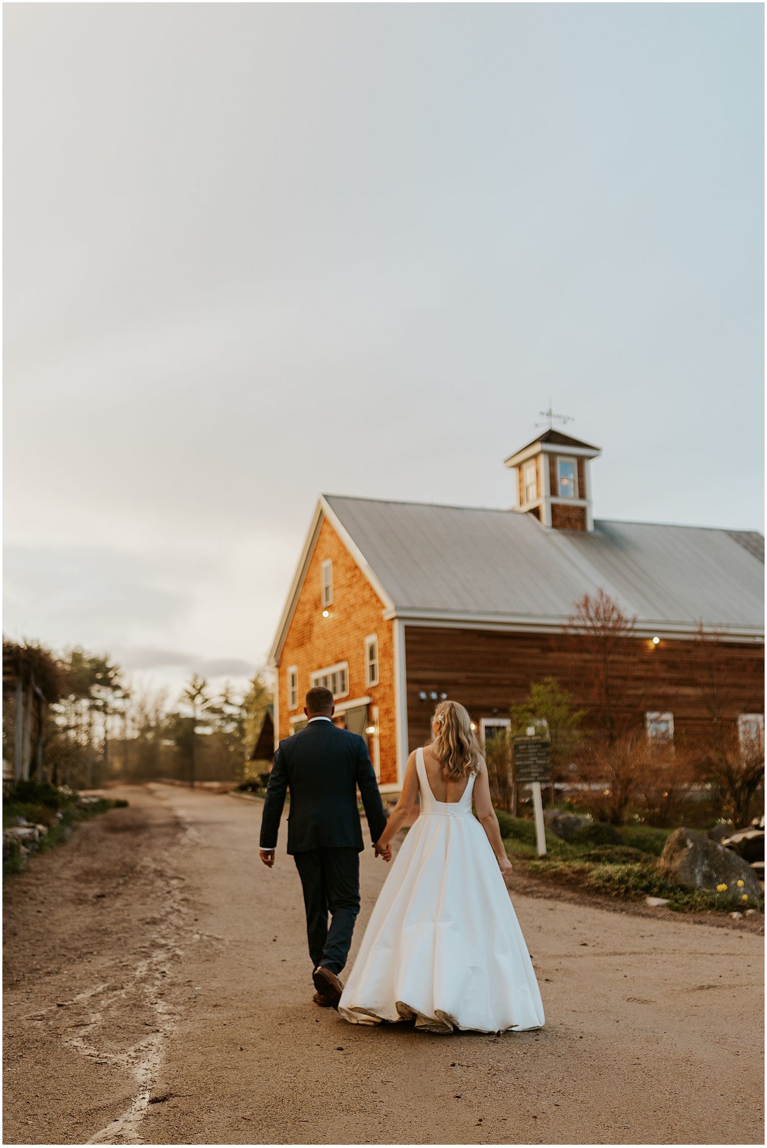 A couple walks along a dirt path towards a rustic brick church building at sunset.