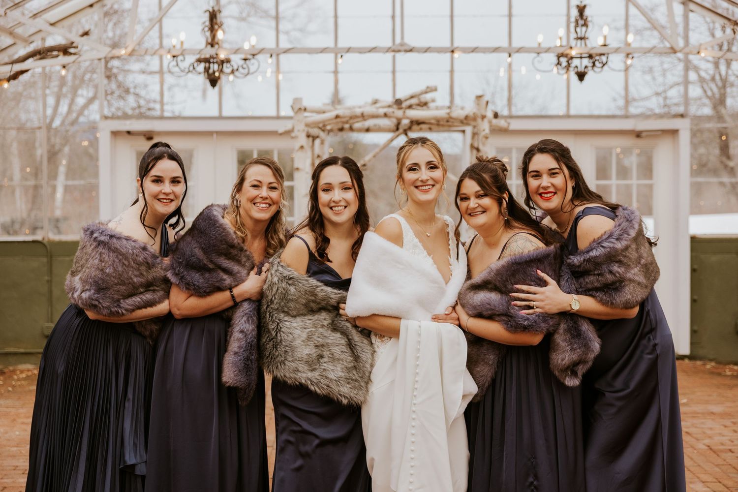 Bride and five bridesmaids in navy dresses with fur stoles posing in front of greenhouse.