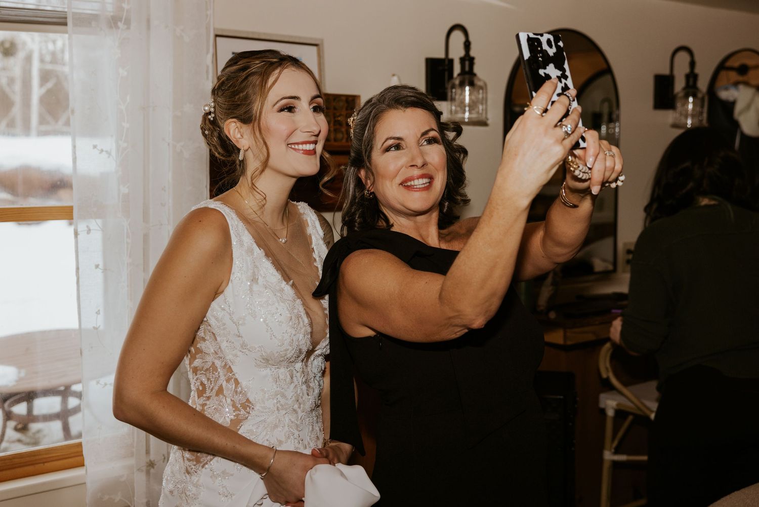 Bride in lace wedding dress takes a selfie with a woman in black at an indoor wedding venue.