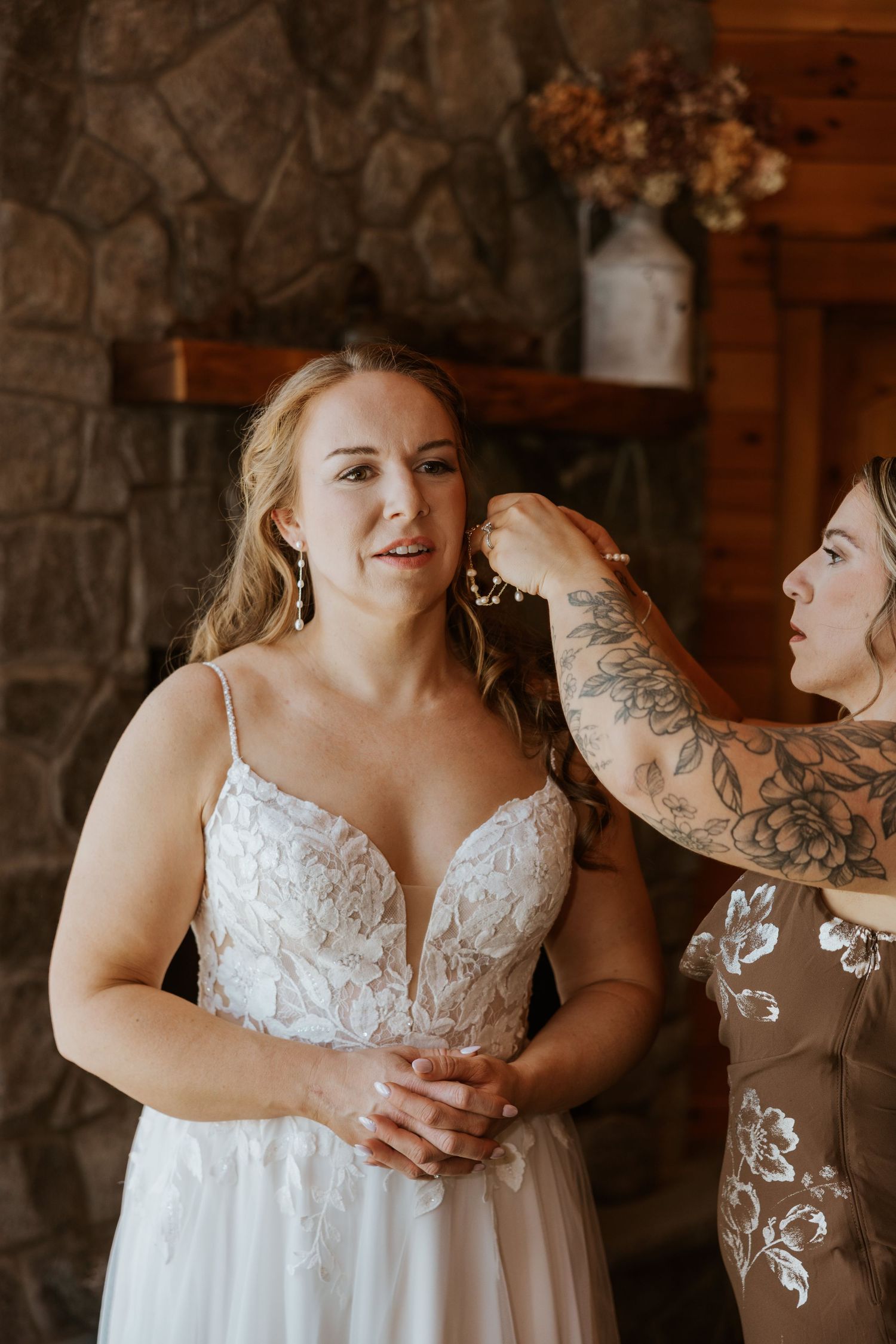 A wedding dress fitting moment captured against a stone fireplace in a rustic cabin setting.