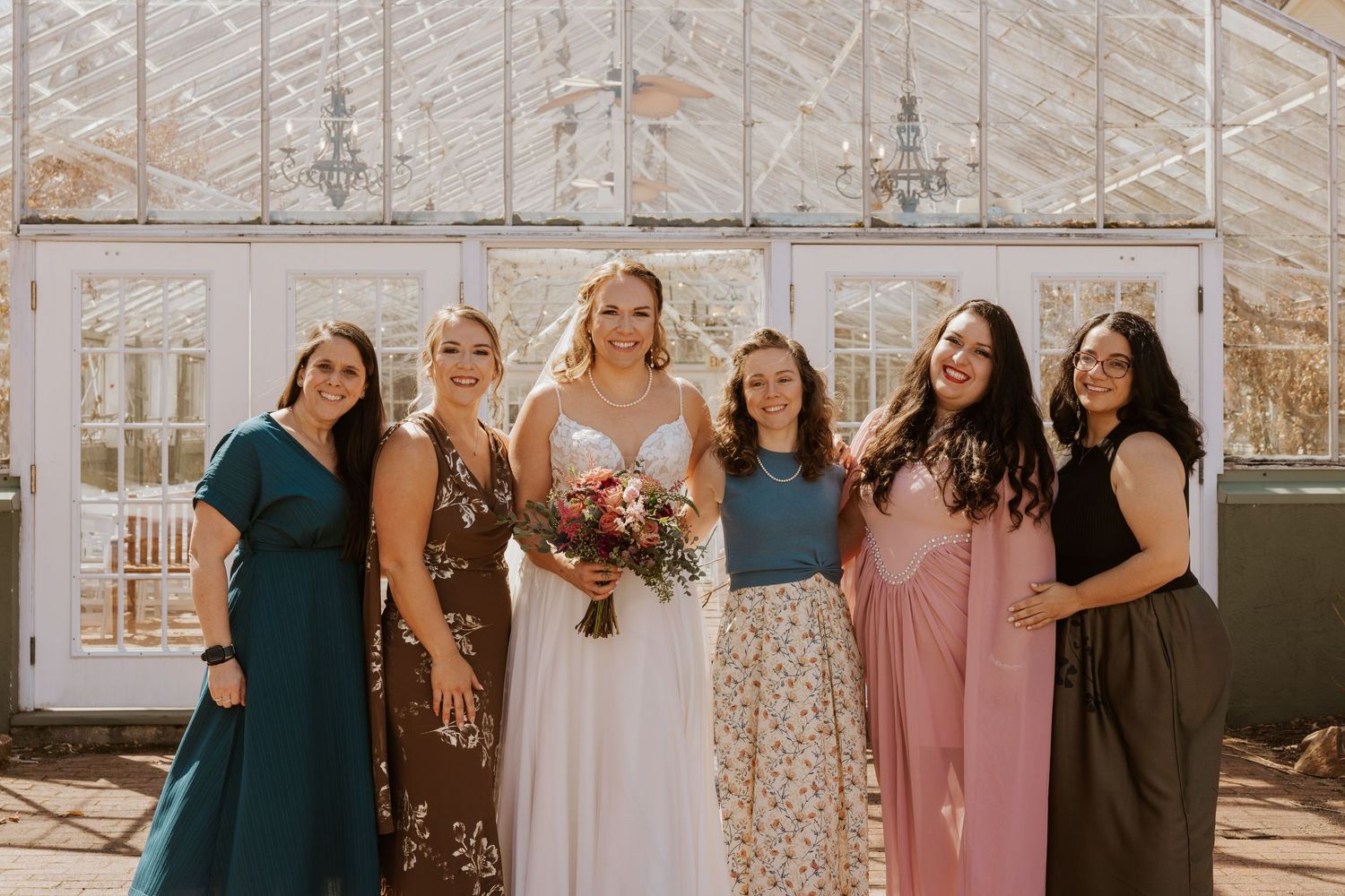 A wedding party in coordinated dresses stands together in a greenhouse with natural light streaming through glass walls.