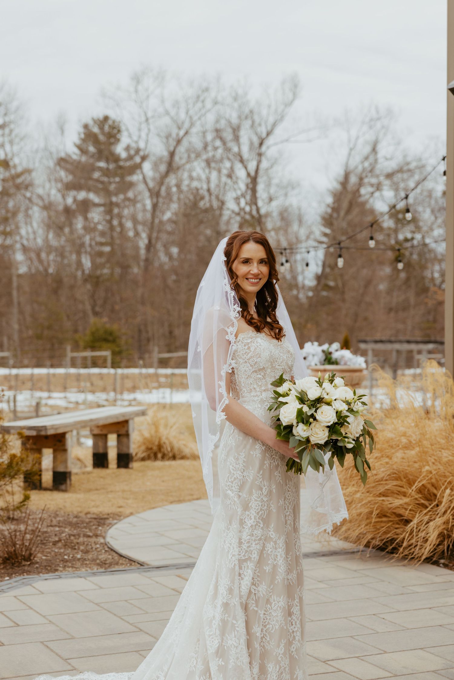 A bride in a lace wedding dress holds a white floral bouquet while standing on a stone path surrounded by winter foliage.