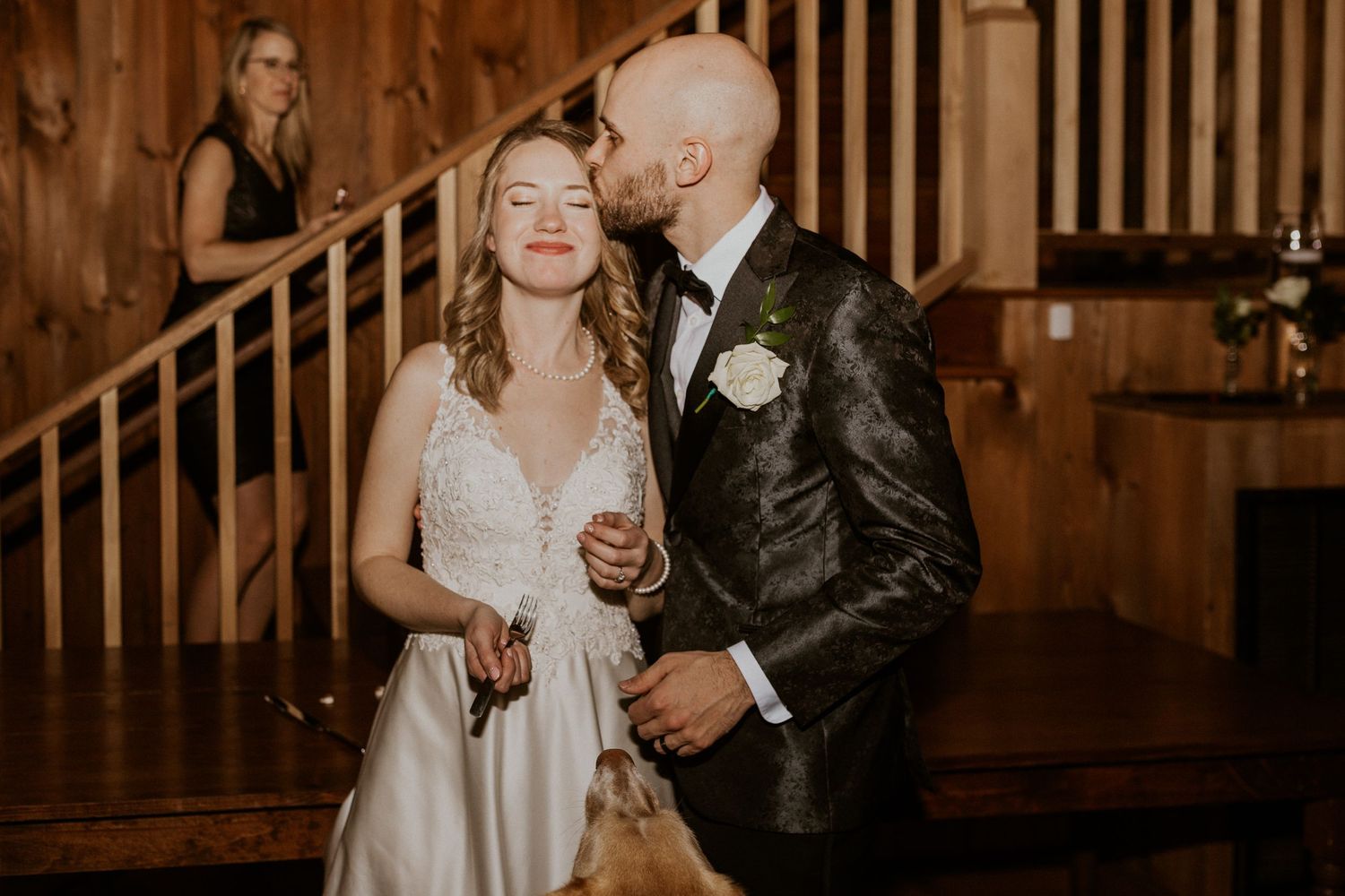Brodie kissing Deirdre on the temple after they cut their wedding cake, their pup Kenobi is in the foreground.