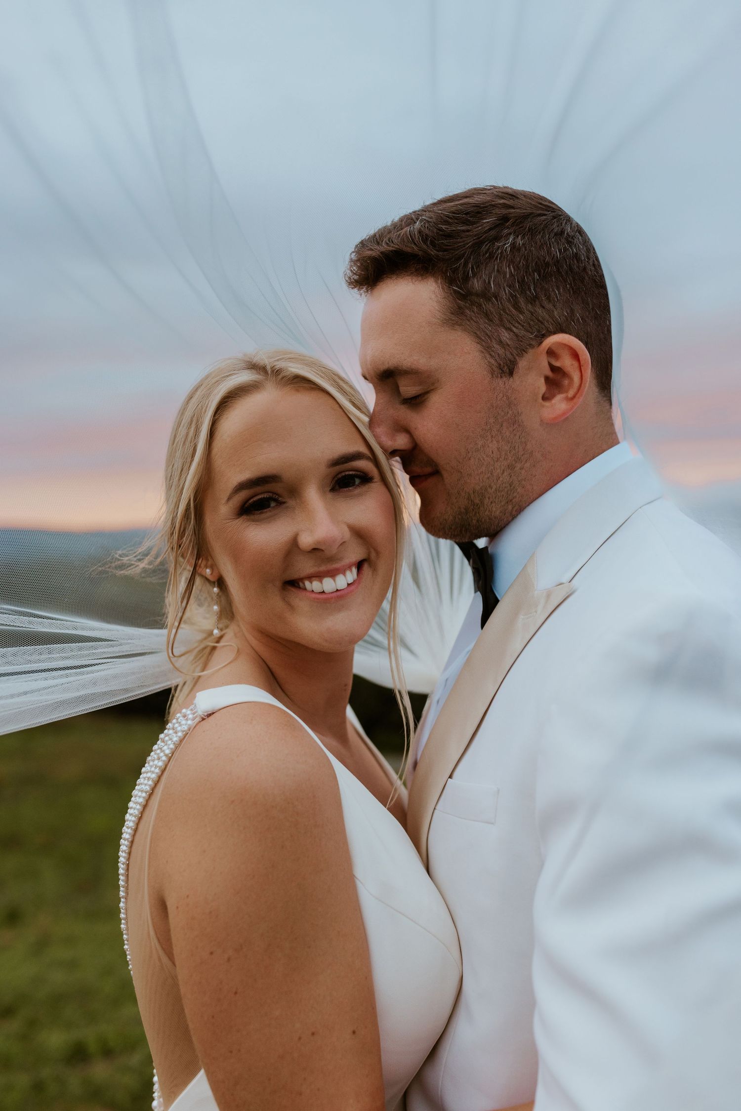 Romantic wedding couple embracing outdoors at sunset with flowing veil.