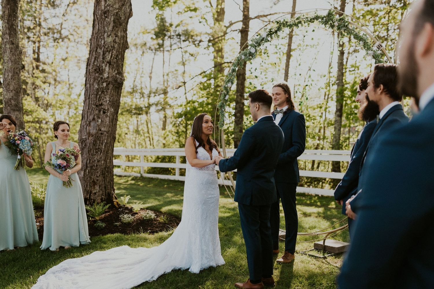 Chrissy and Mike holding hands as their wedding ceremony begins. It is a lovely spring day with the sun shining.