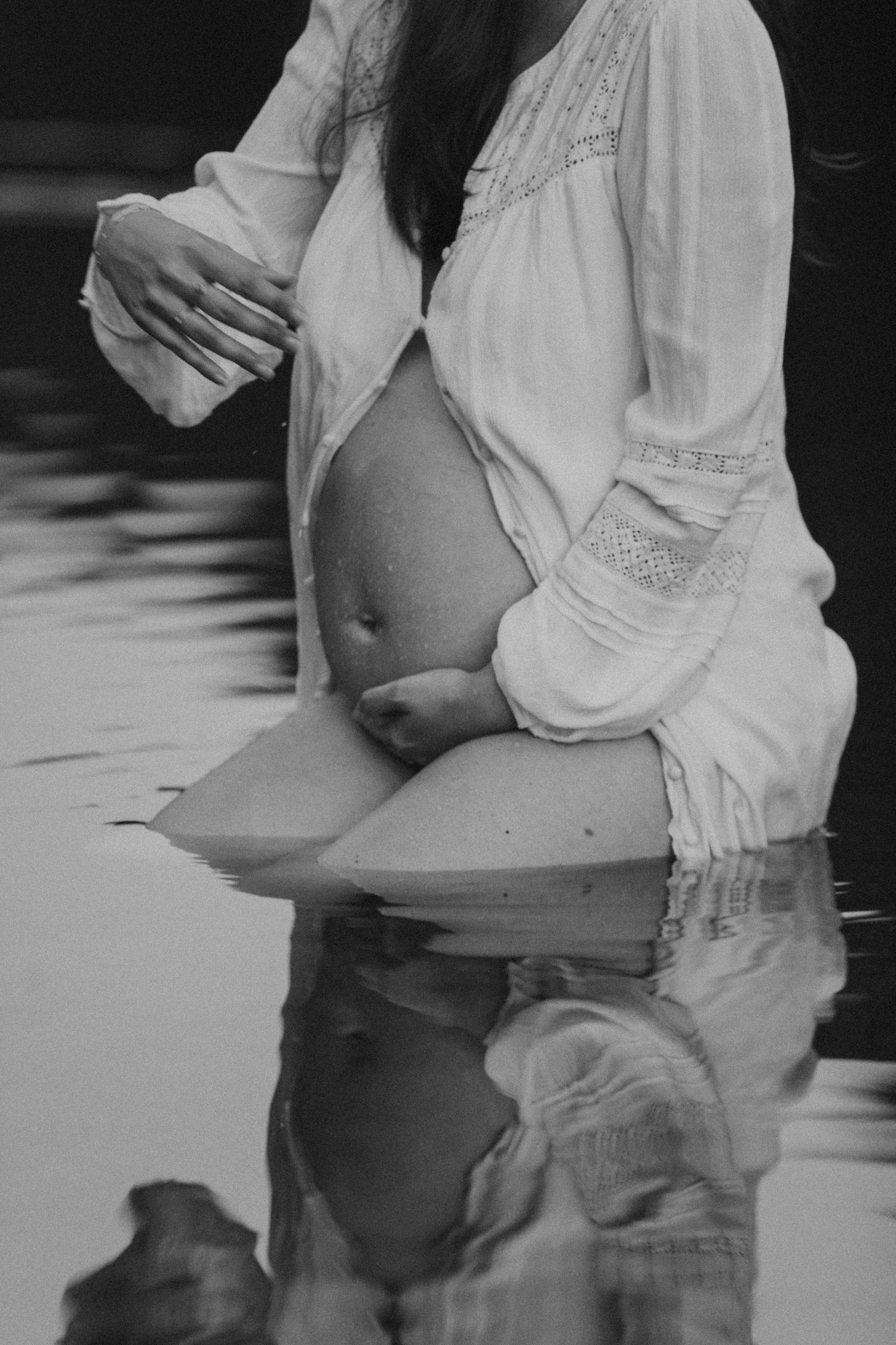 Black and white maternity photo of a pregnant woman cradling her bare belly while standing in water.