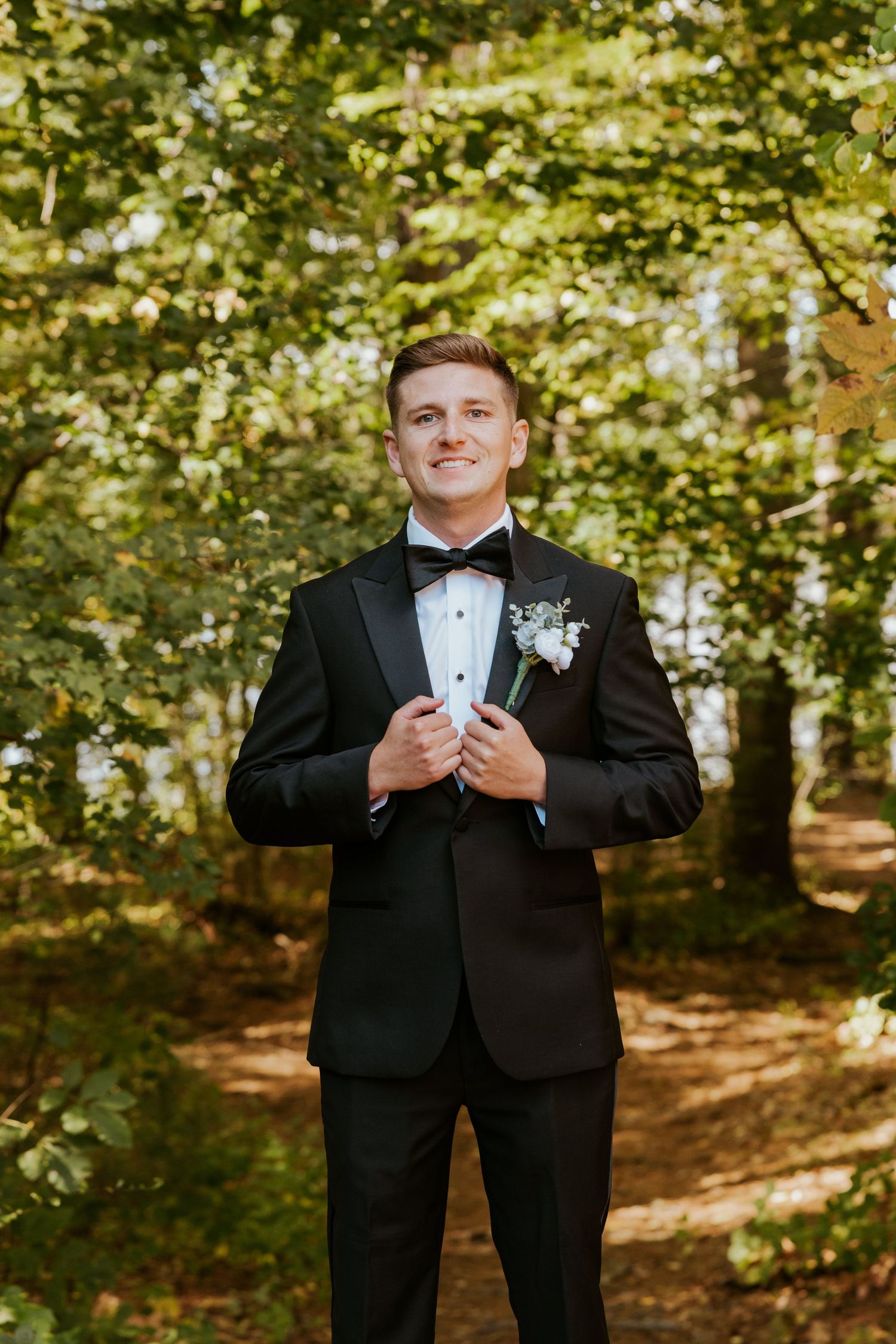 Handsome groom in black tuxedo with boutonniere smiles confidently in sunlit forest setting.