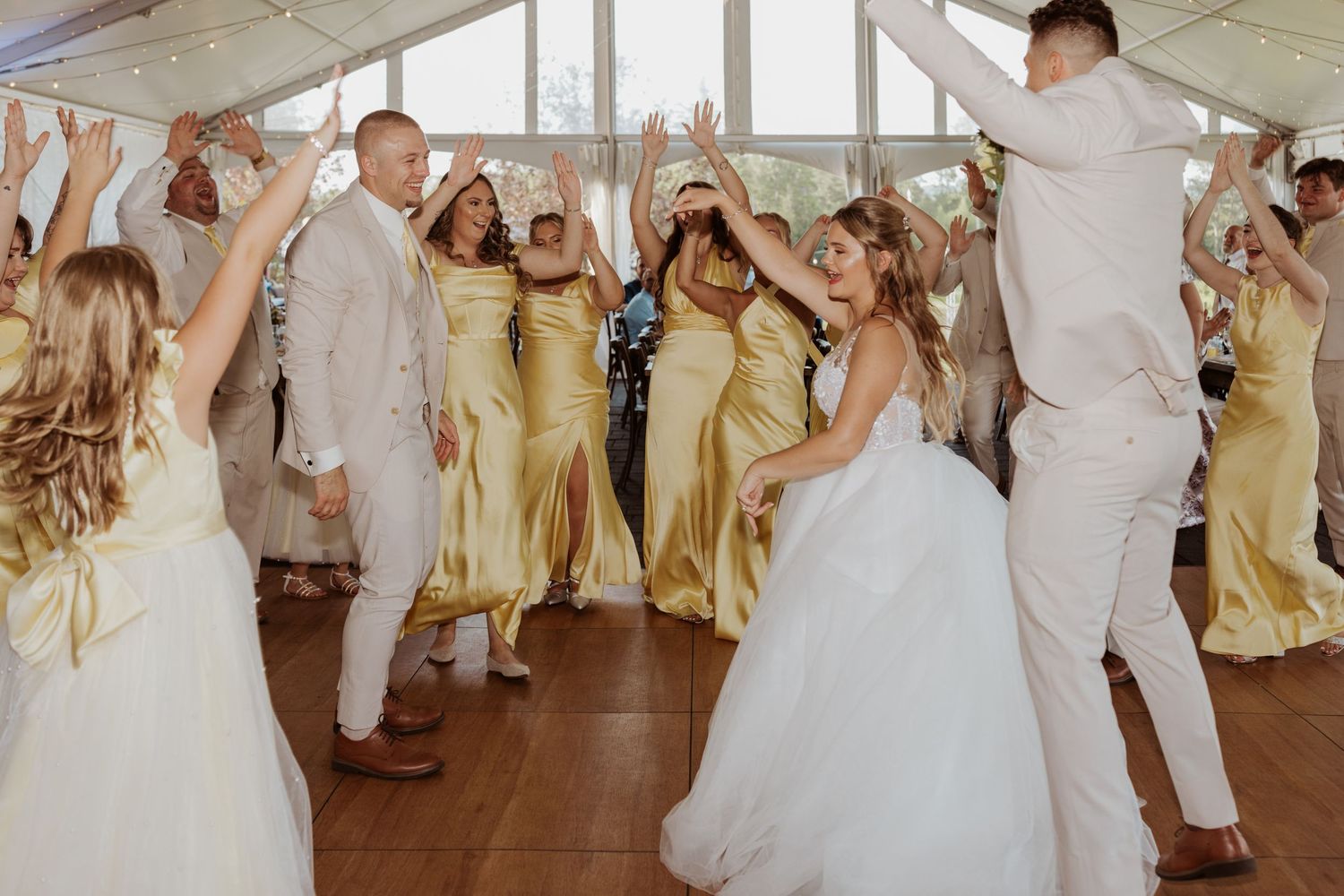 Bride, groom, and gold-dressed bridesmaids dancing joyfully at wedding reception under tent.