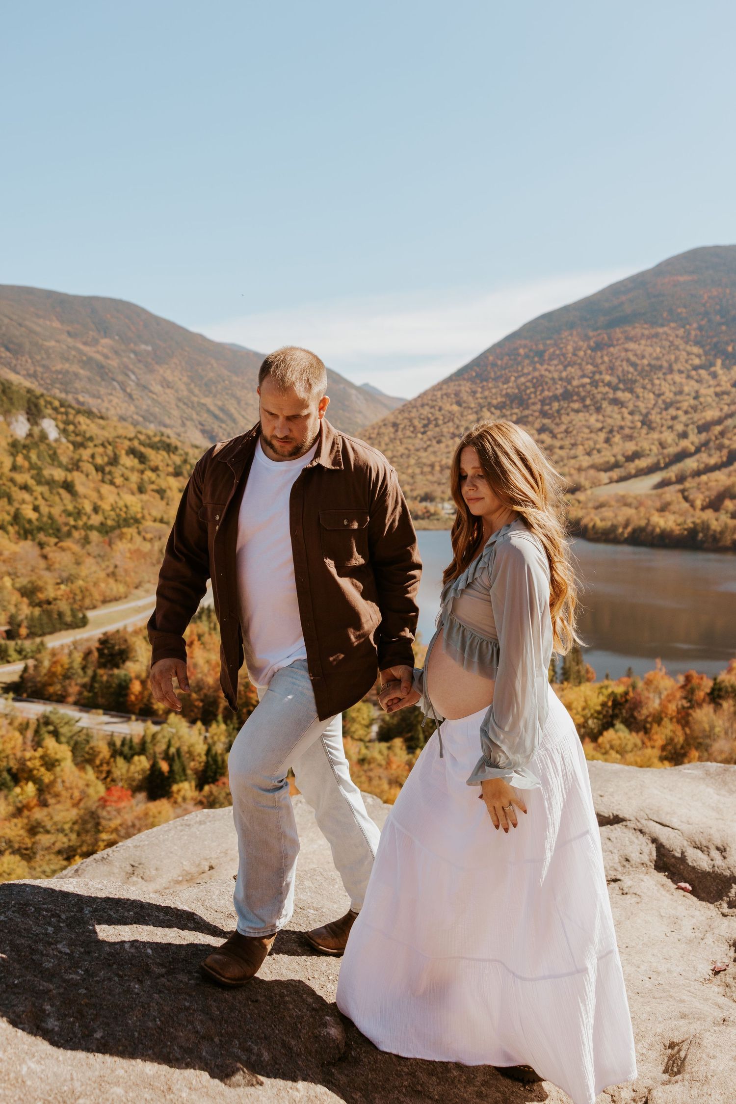 Couple walking hand-in-hand on mountain summit with vibrant autumn lake and hillside scenery below.