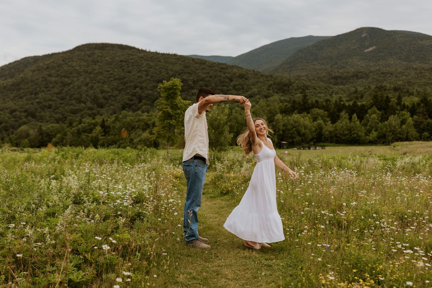 Joyful couple dancing and twirling in a wildflower meadow with scenic mountain landscape behind.