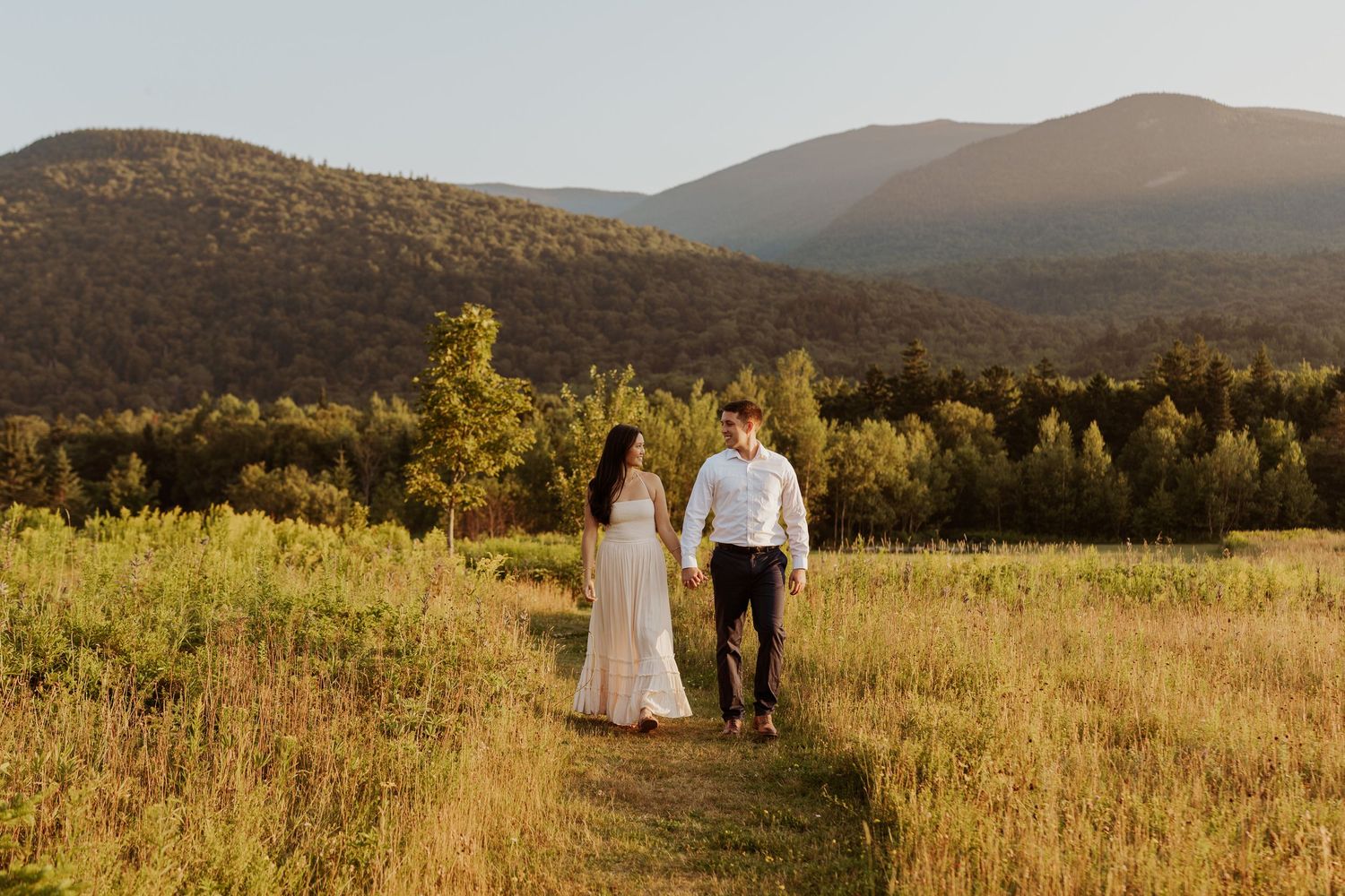 Couple walks hand in hand through golden meadow with mountain backdrop at sunset.