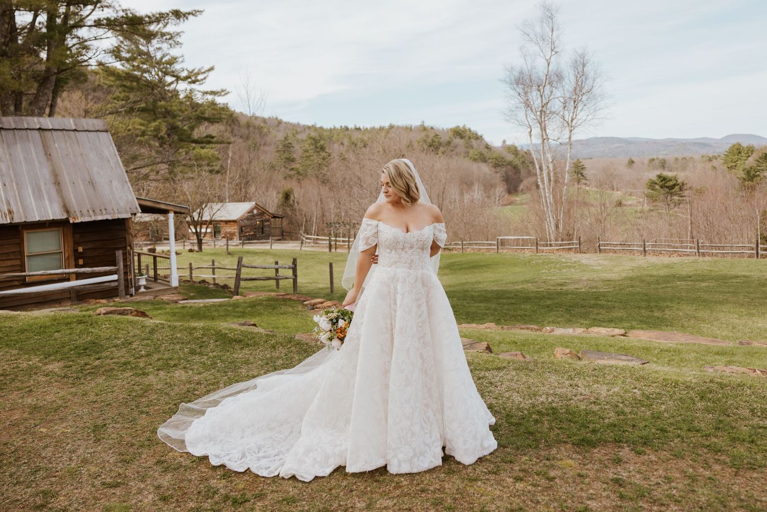 Bride in off-shoulder lace ball gown with long train standing in a rustic farm landscape.