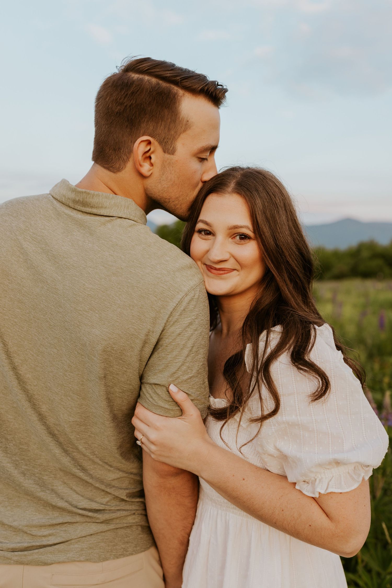 Man kissing woman on the cheek in a lupine field, bride smiling at camera during engagement shoot.