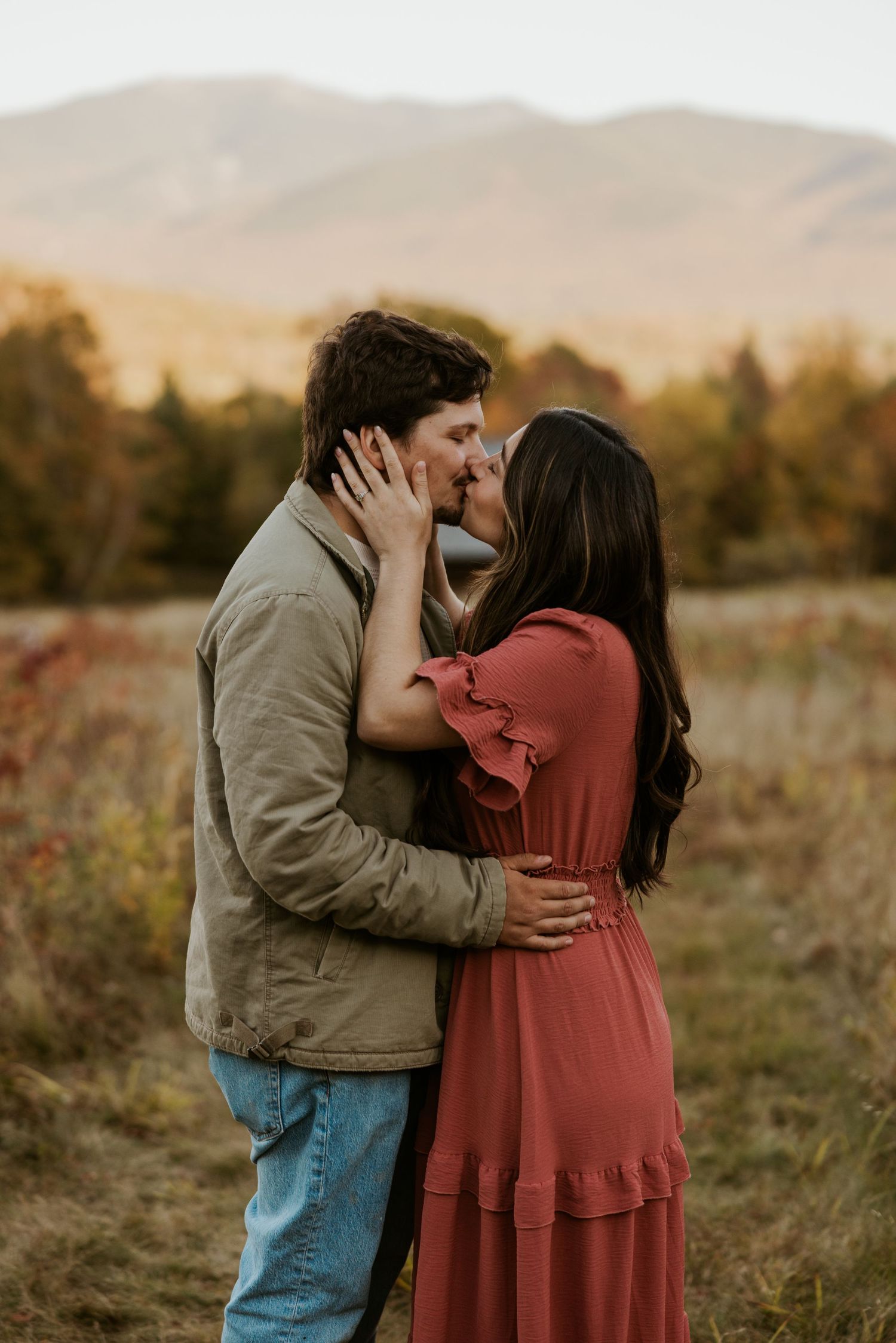 Couple sharing a passionate kiss in an open field with warm golden autumn mountains in the background.